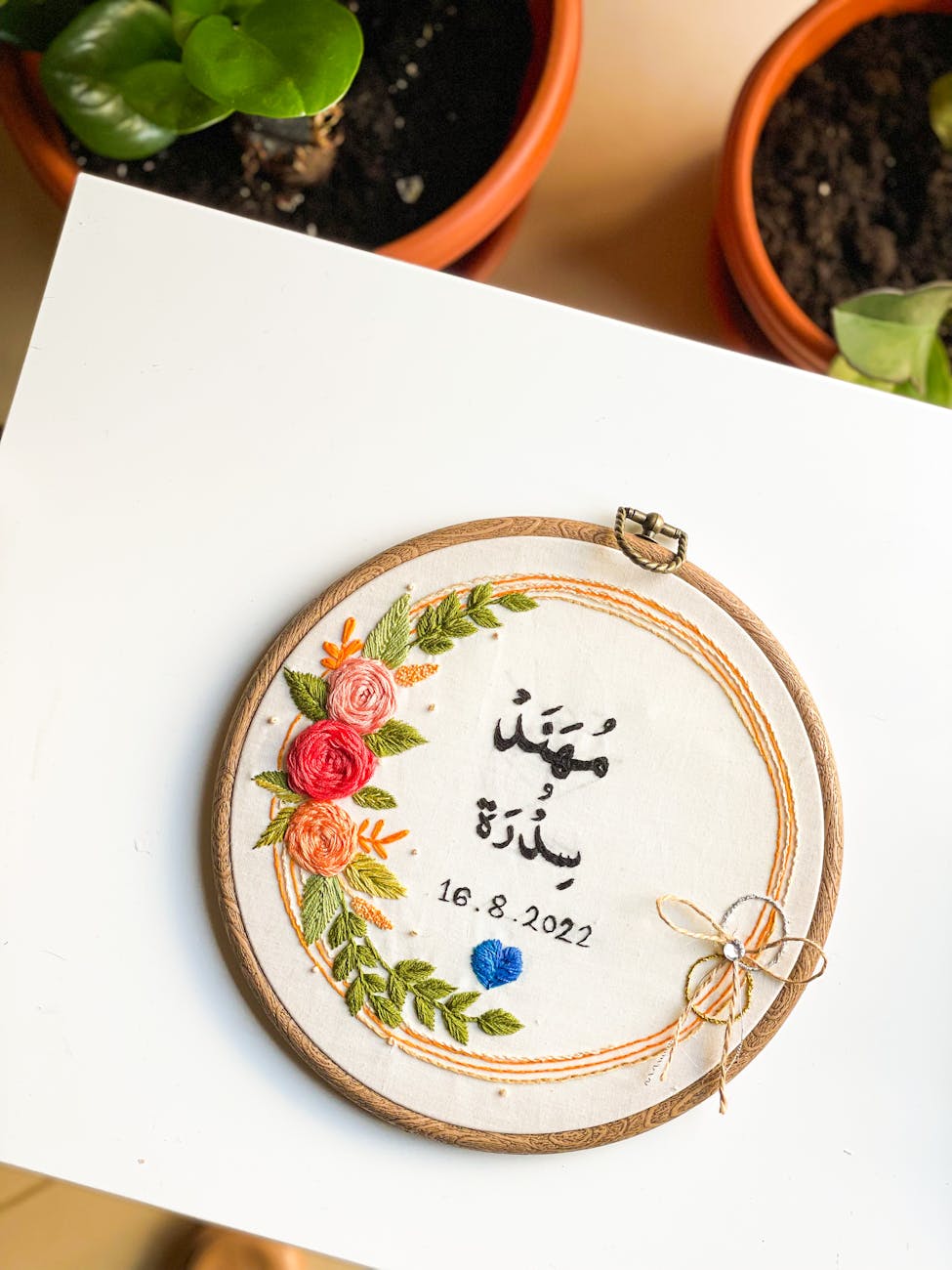 Embroidered wooden hoop featuring Arabic calligraphy names and date "16.8.2022", floral wreath embroidery in pink, orange and green threads, small blue heart and twine bow, placed on white surface with terracotta potted plants.
