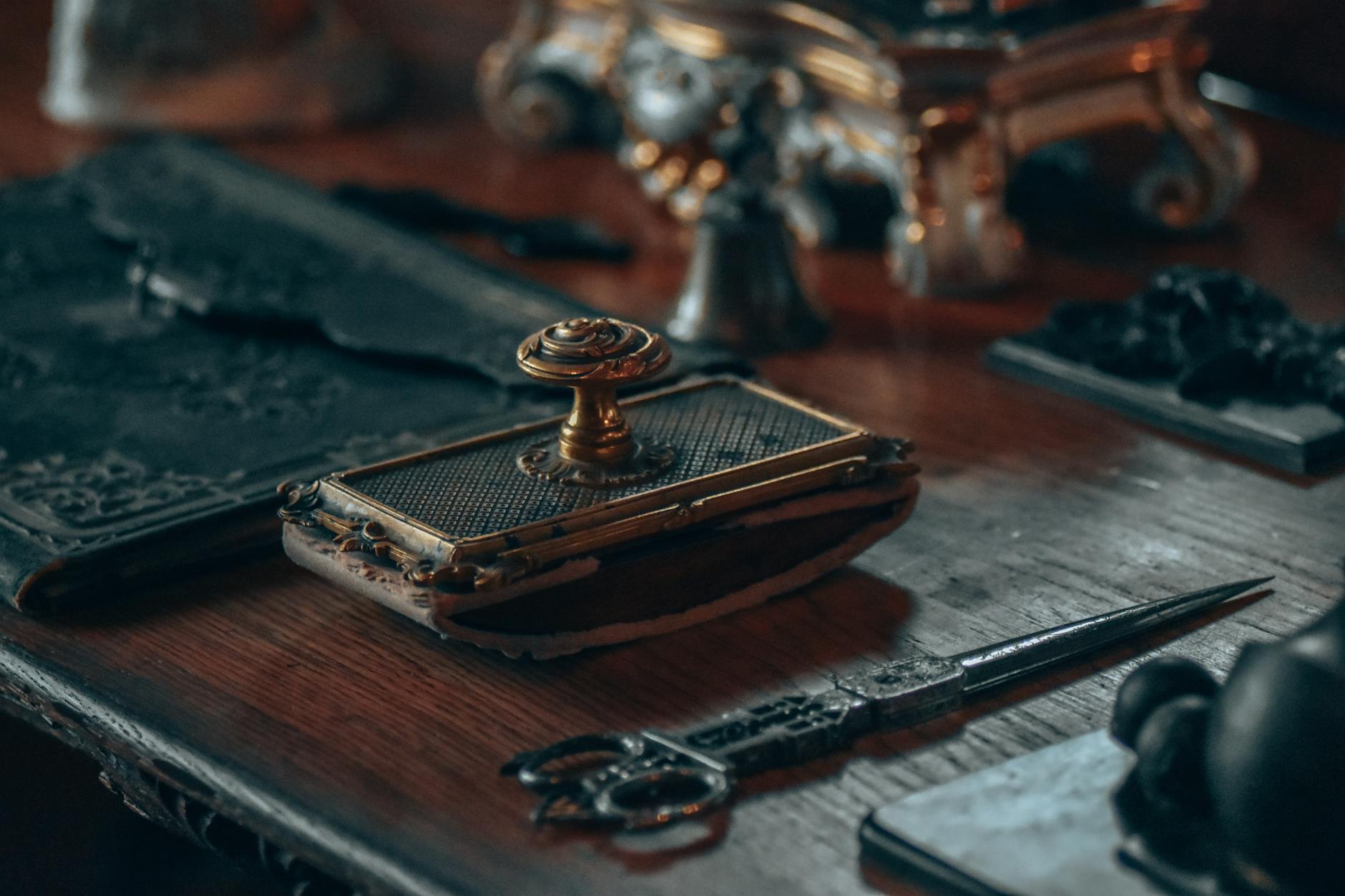 Vintage writing desk with ornate brass wax seal stamp, decorative scissors, letter opener and leather-bound book on polished wood surface