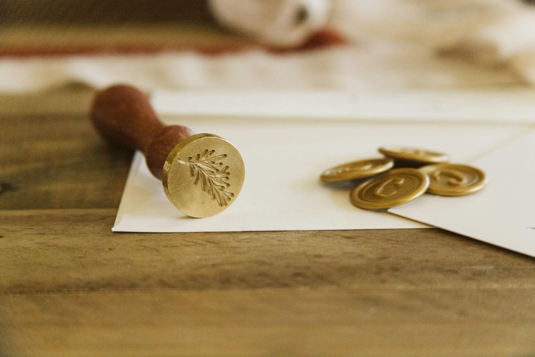 Brass wax seal stamp with wooden handle engraved with a fern design resting on white envelopes beside stacked gold wax seals on a wooden table