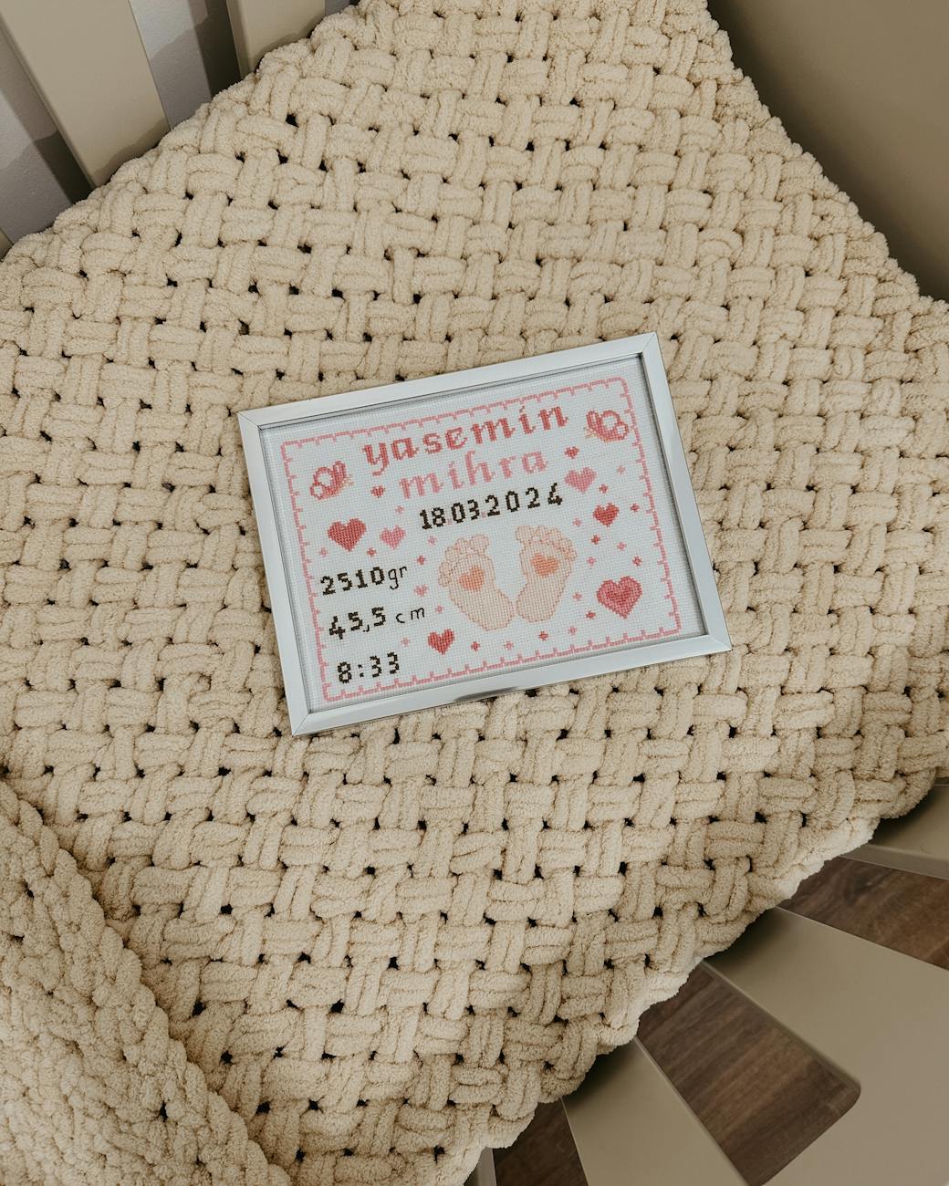 Framed pink cross-stitch birth announcement reading "Yasemin Mihra 18.03.2024" with baby footprints, weight 2510 g, length 45.5 cm and time 8:33, placed on a beige chunky knit blanket inside a baby crib