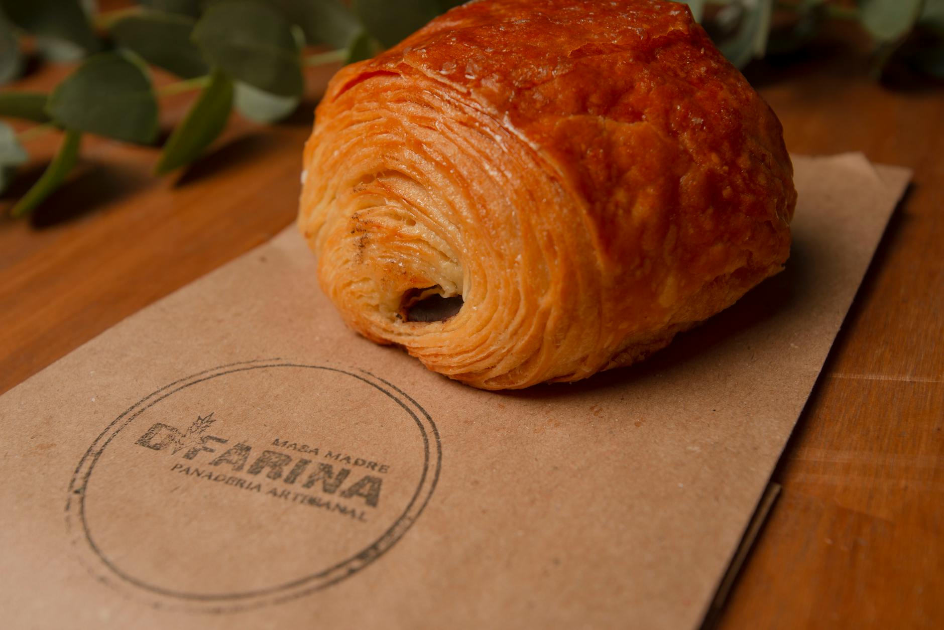 Buttery flaky pain au chocolat (chocolate croissant) on branded kraft bakery paper, close-up showing layers and golden brown crust on wooden table