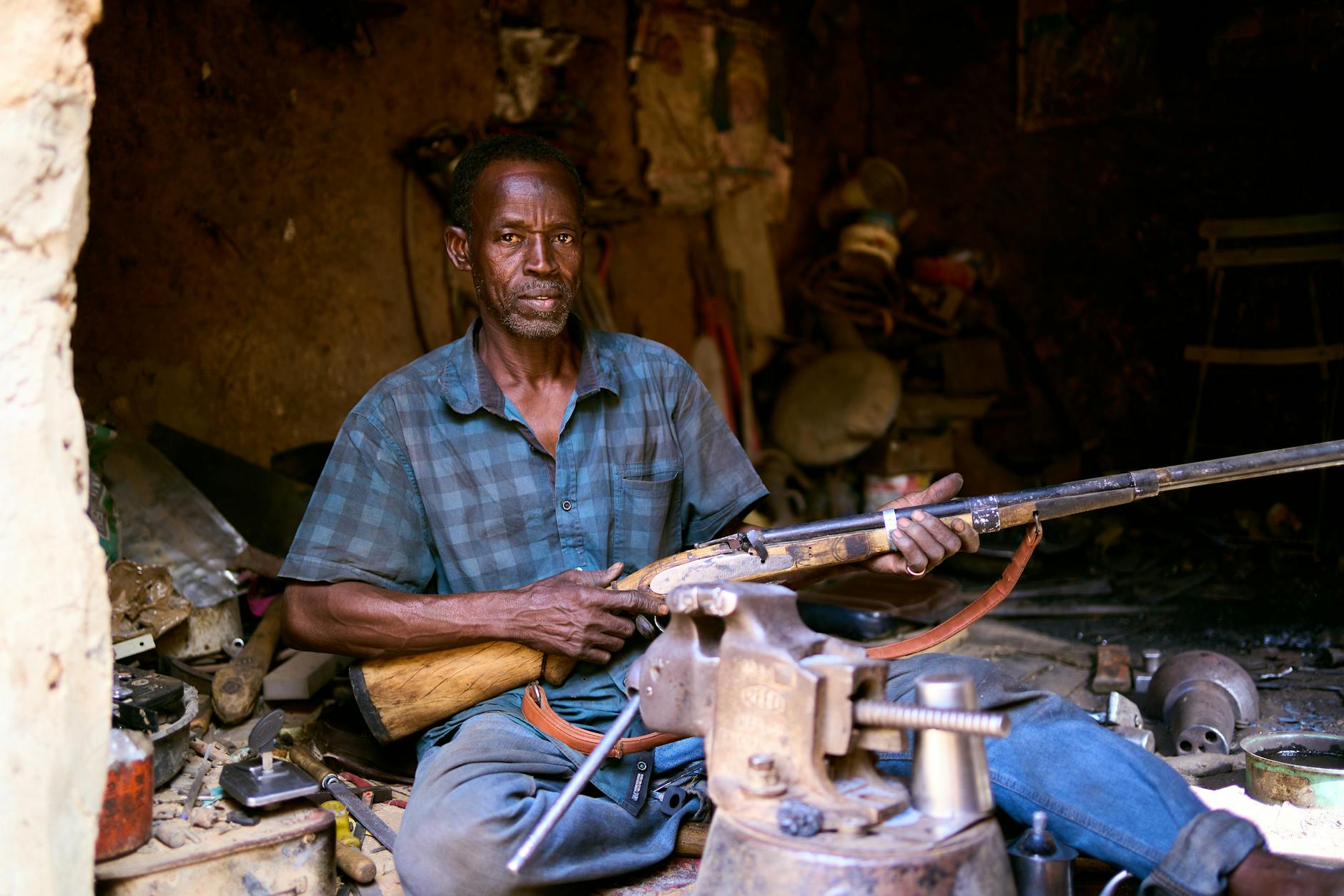 Seated man in a rustic workshop holding a handcrafted rifle, surrounded by metalworking tools, a bench vise, spare parts and a cluttered workbench—traditional gunsmith repair scene in a dim, earthy interior