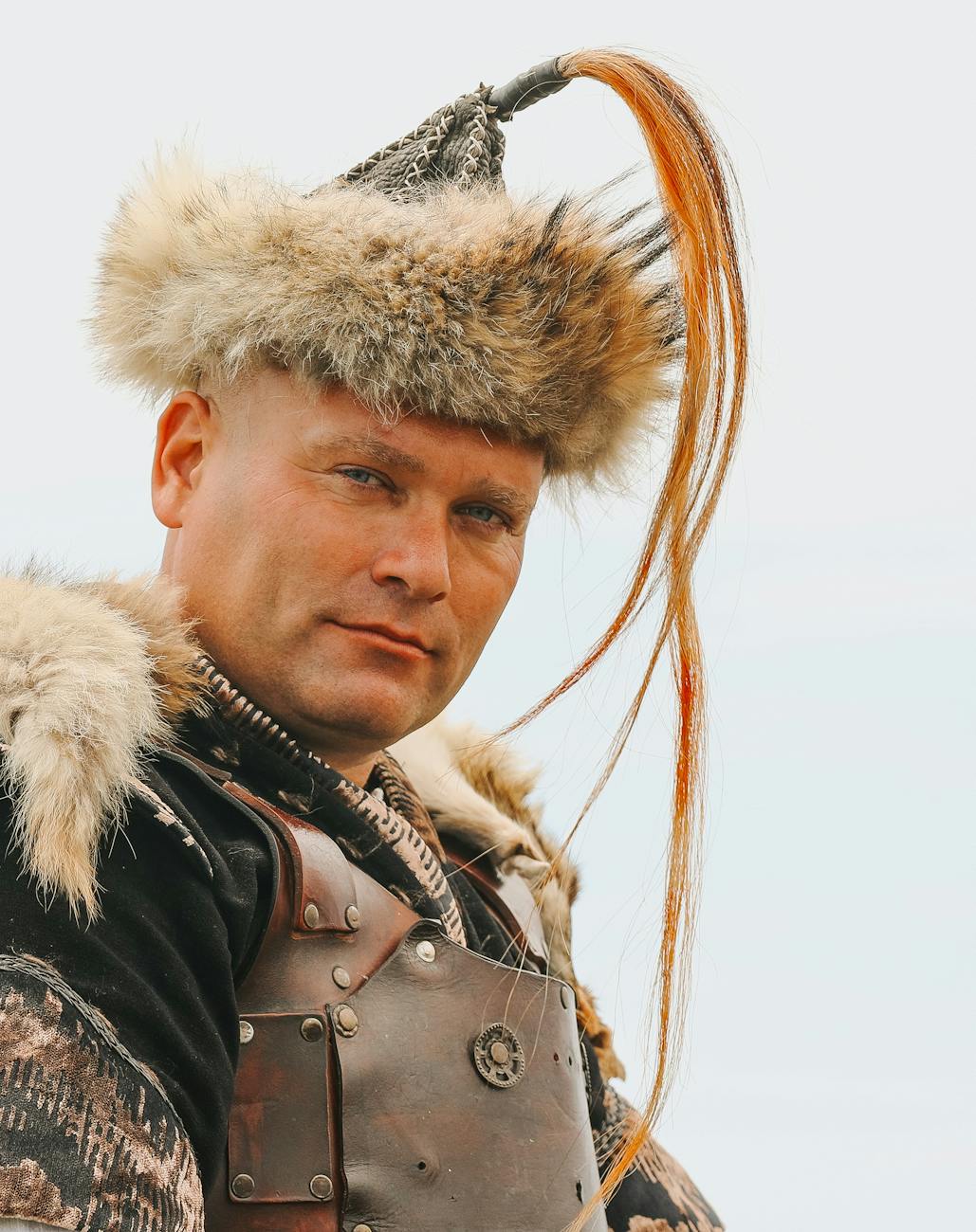 Portrait of a Viking-style reenactor wearing a fur hat with a long horsehair plume, leather armor and fur cloak, stoic expression and weathered outdoor look