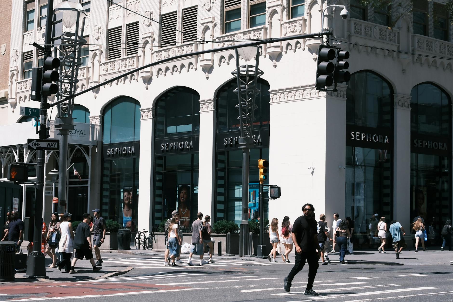 Sephora storefront on a busy city corner with ornate white facade, pedestrians crossing at a traffic light on 5th Avenue, urban shopping scene