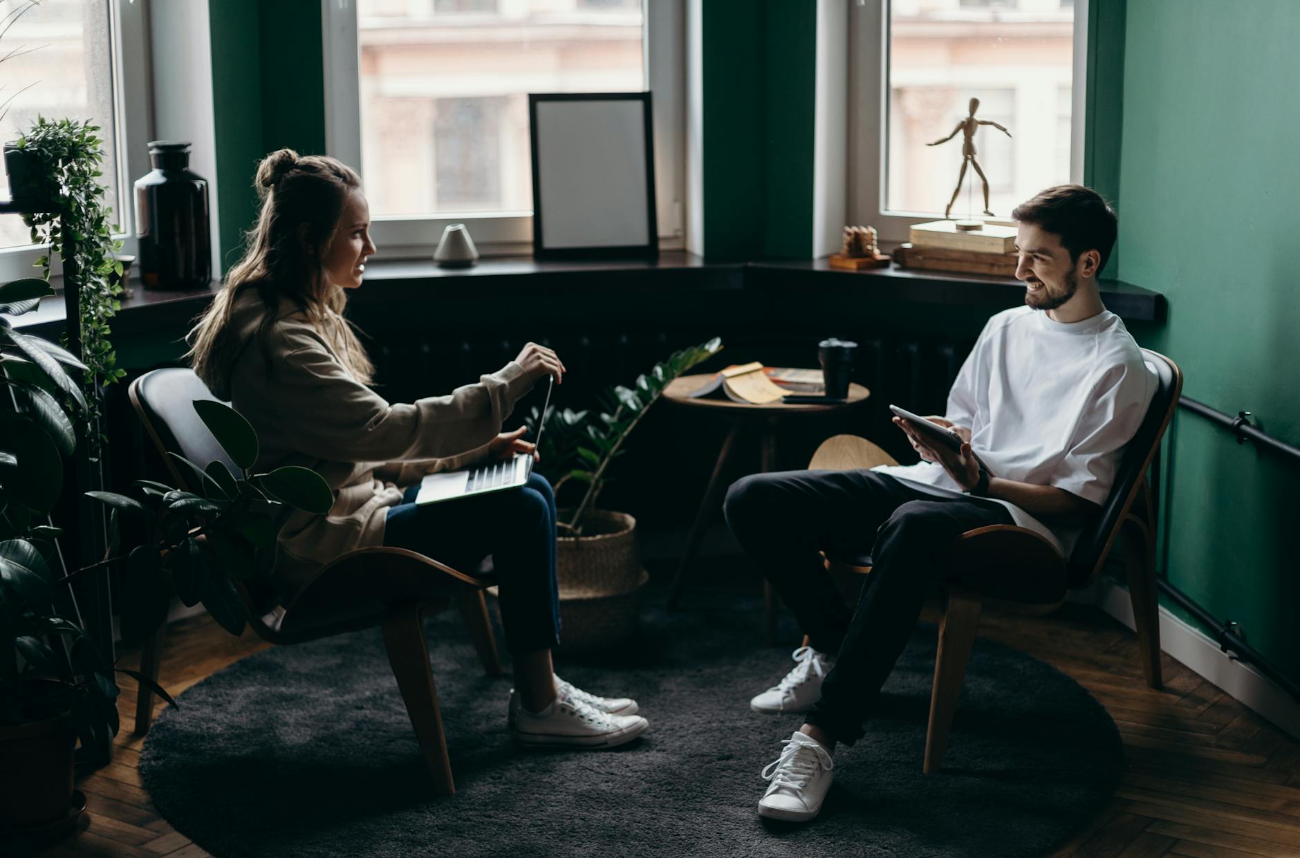 Two people having a relaxed meeting in a cozy green office lounge with laptops and tablet, indoor plants, natural light and modern wooden chairs