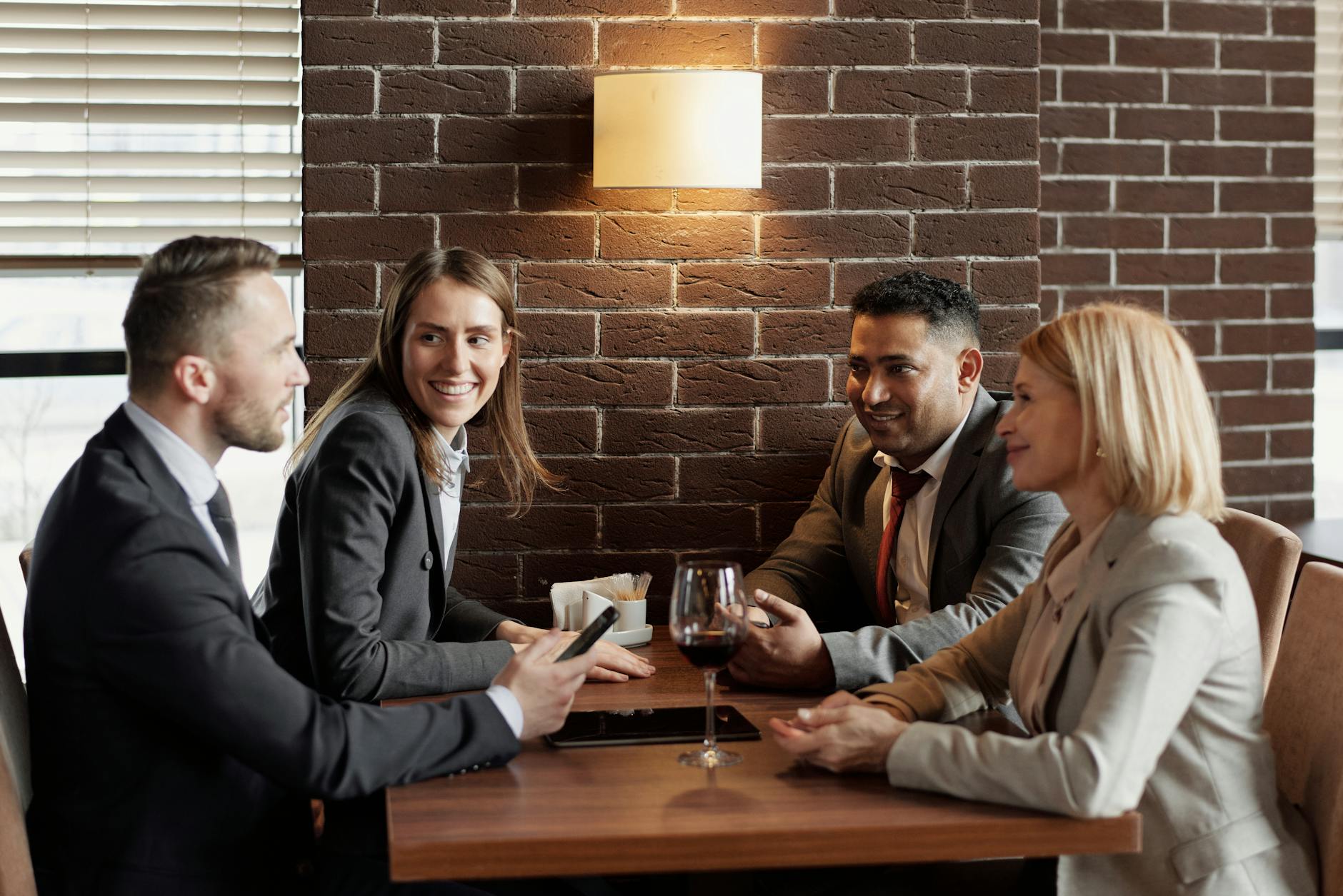 Four business professionals in suits having a relaxed meeting at a wooden table in a brick-walled restaurant, wine glass and tablet visible