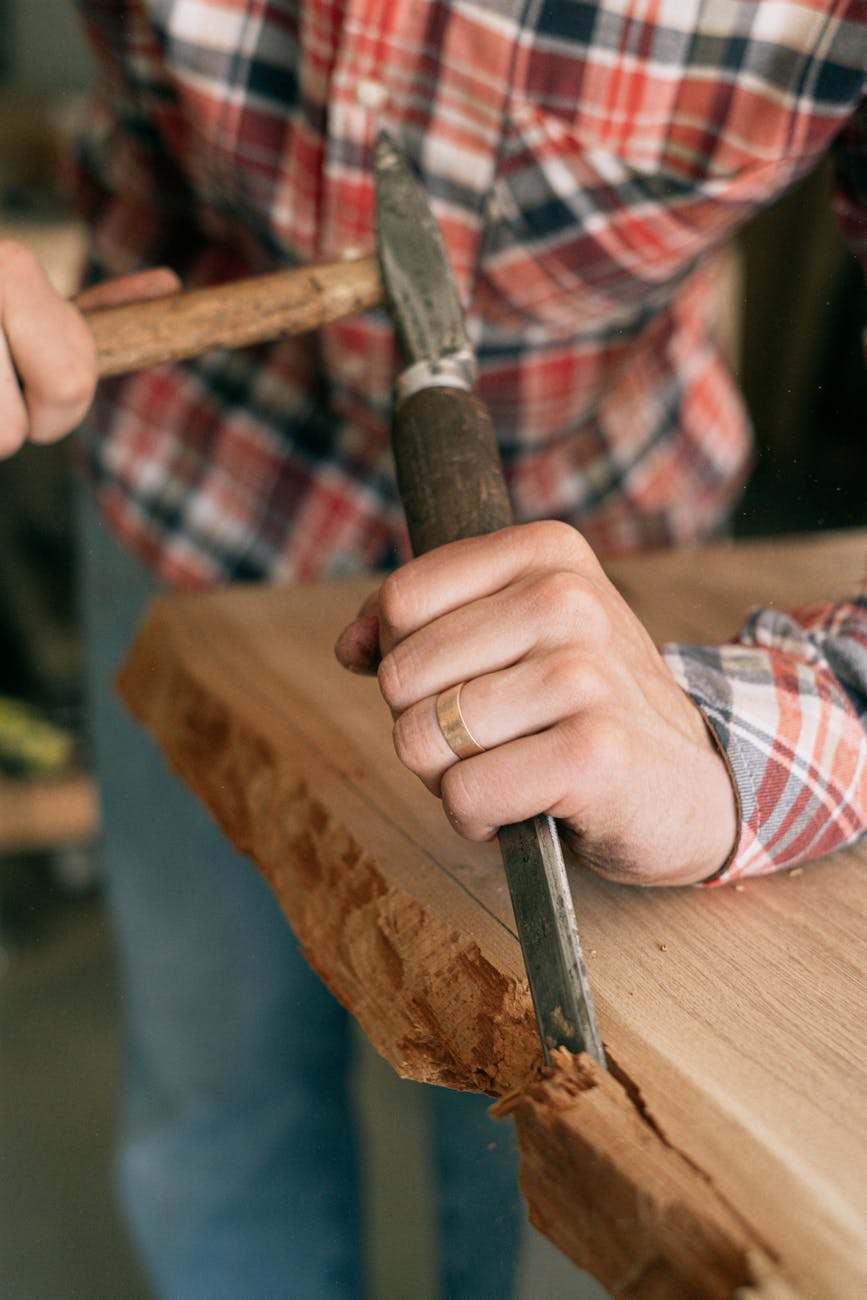 Close-up of a woodworker chiseling a live-edge oak slab with a mallet and chisel, hands and woodgrain in focus, rustic woodworking tools and flannel shirt.