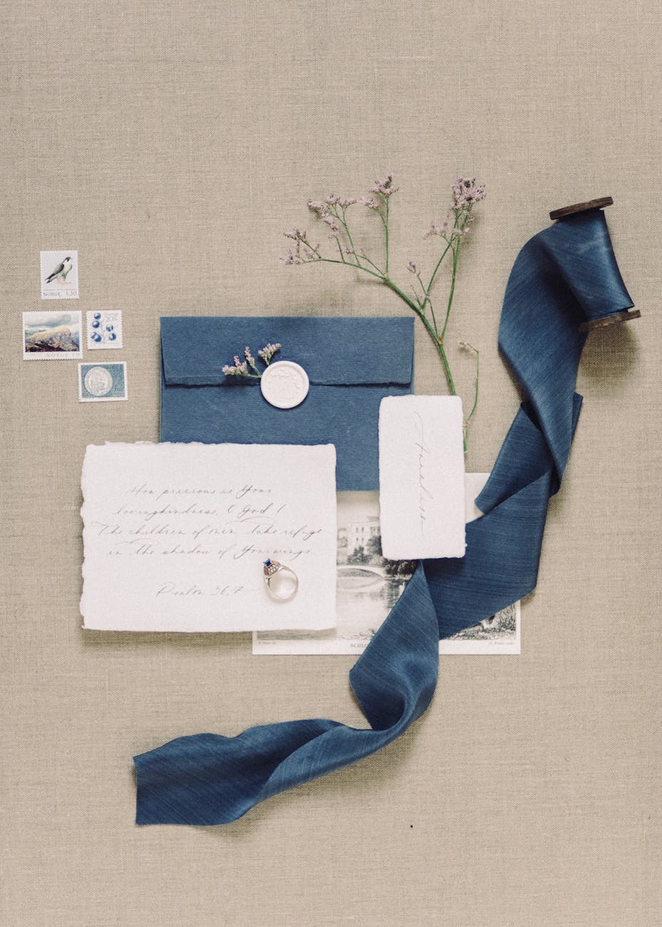 Wedding invitation flat lay with navy envelope sealed with wax, handwritten vow card, sapphire engagement ring, vintage stamps, delicate flowers and flowing blue silk ribbon.