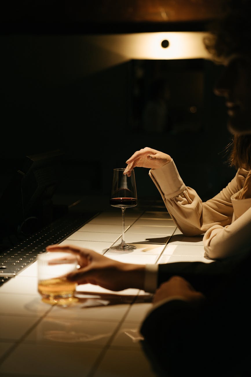 Dimly lit bar counter with two patrons' hands—one holding a whiskey tumbler, the other lightly touching a red wine glass—soft warm lighting and white tiled surface creating an intimate evening atmosphere.