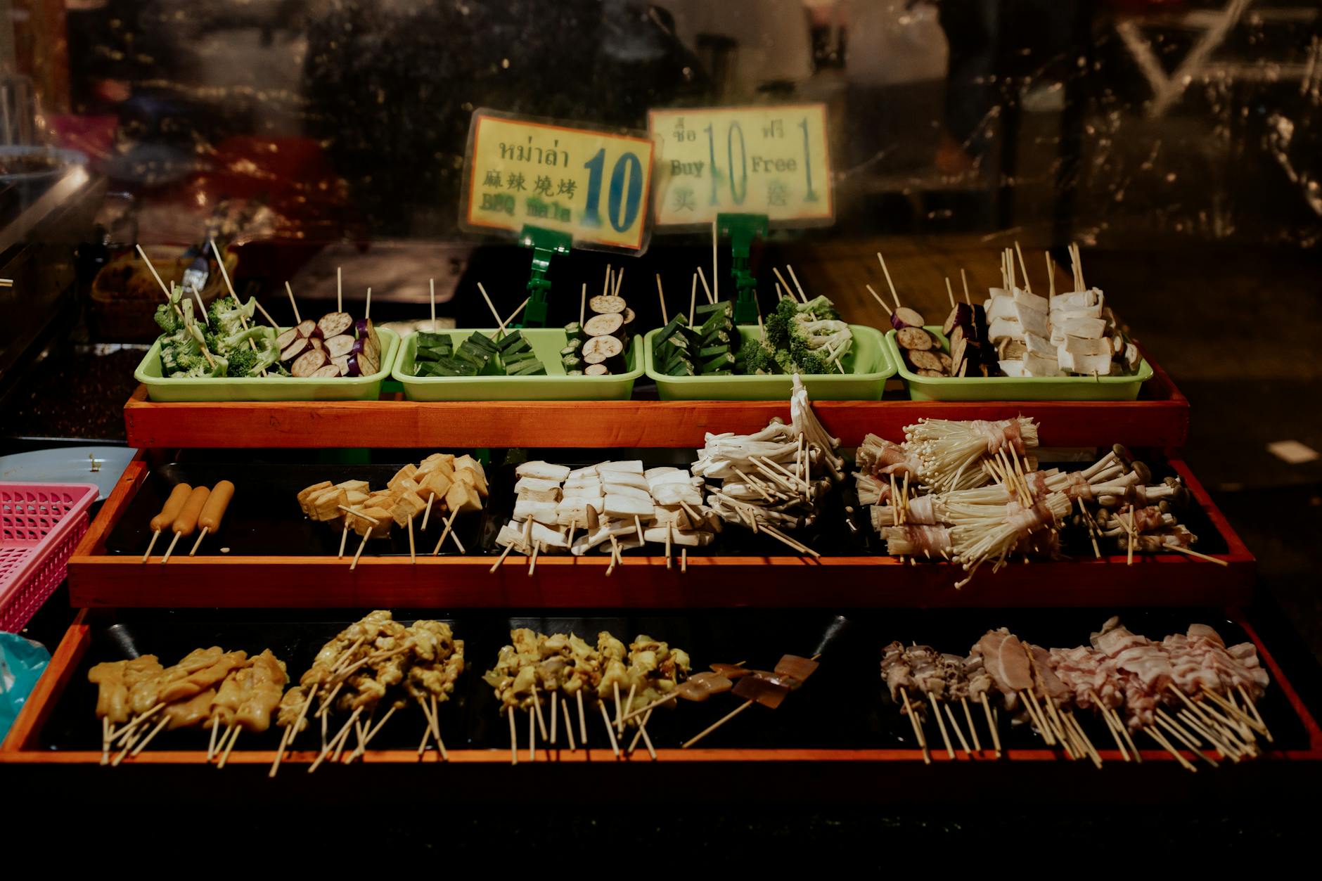 Assorted grilled skewers and vegetables (mushrooms, broccoli, tofu, eggplant) displayed on trays at a Thai street food market stall with price signs.