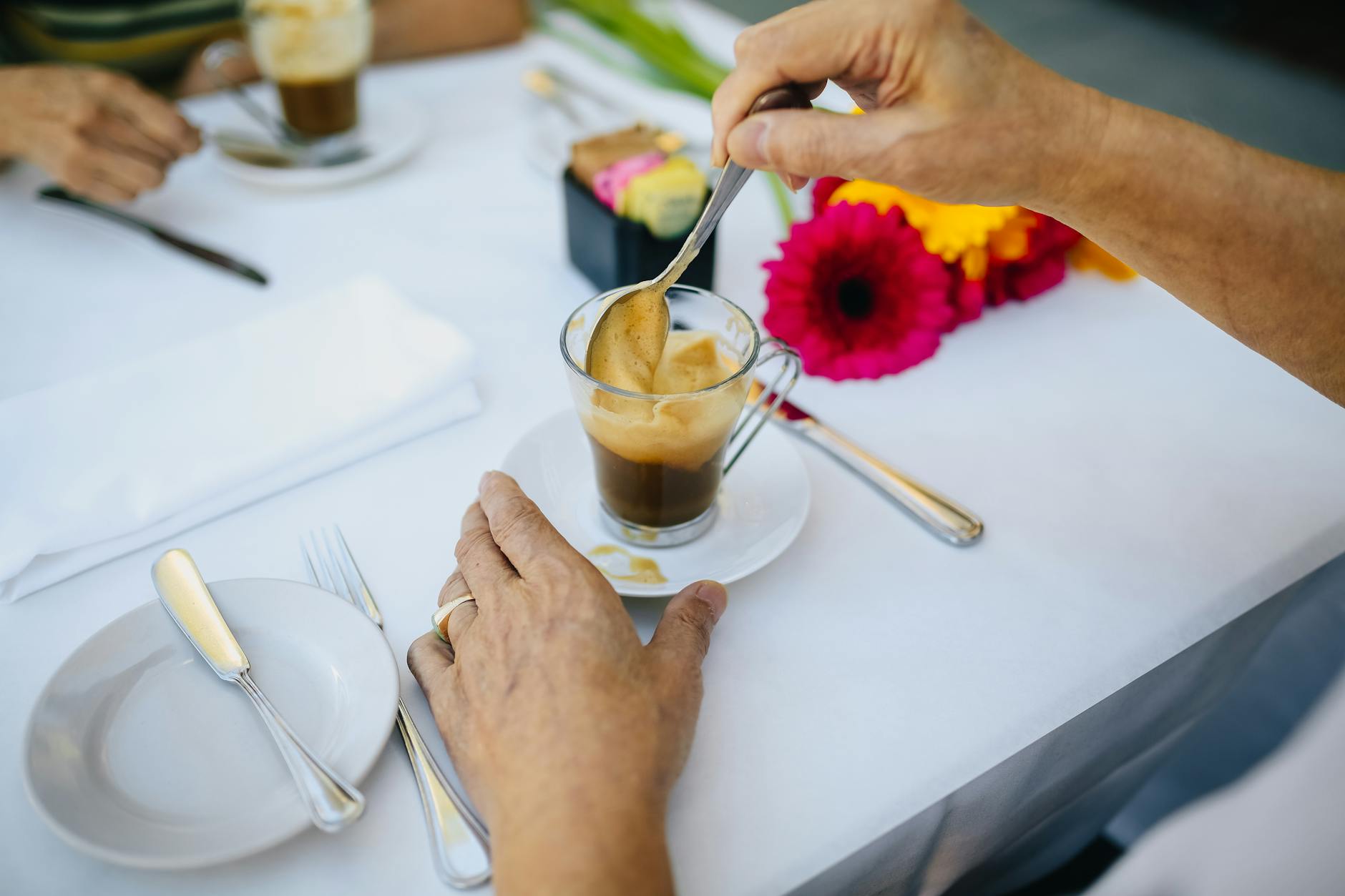 Hands stirring creamy espresso in a glass cup with a spoon on a white cafe table setting, plated saucer, cutlery and bright gerbera flowers.