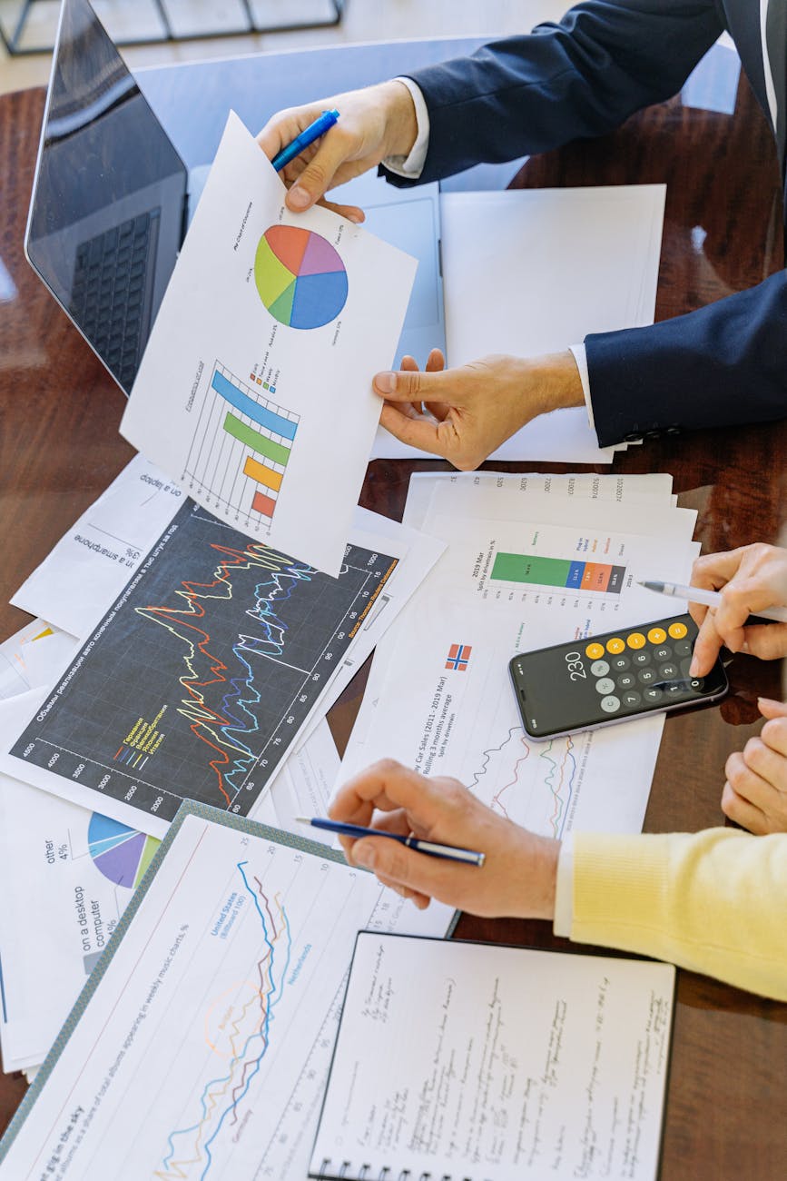 Business team analyzing charts and graphs at a conference table: pie chart, bar chart, line graphs, laptop, smartphone calculator and hands pointing with pens during financial review
