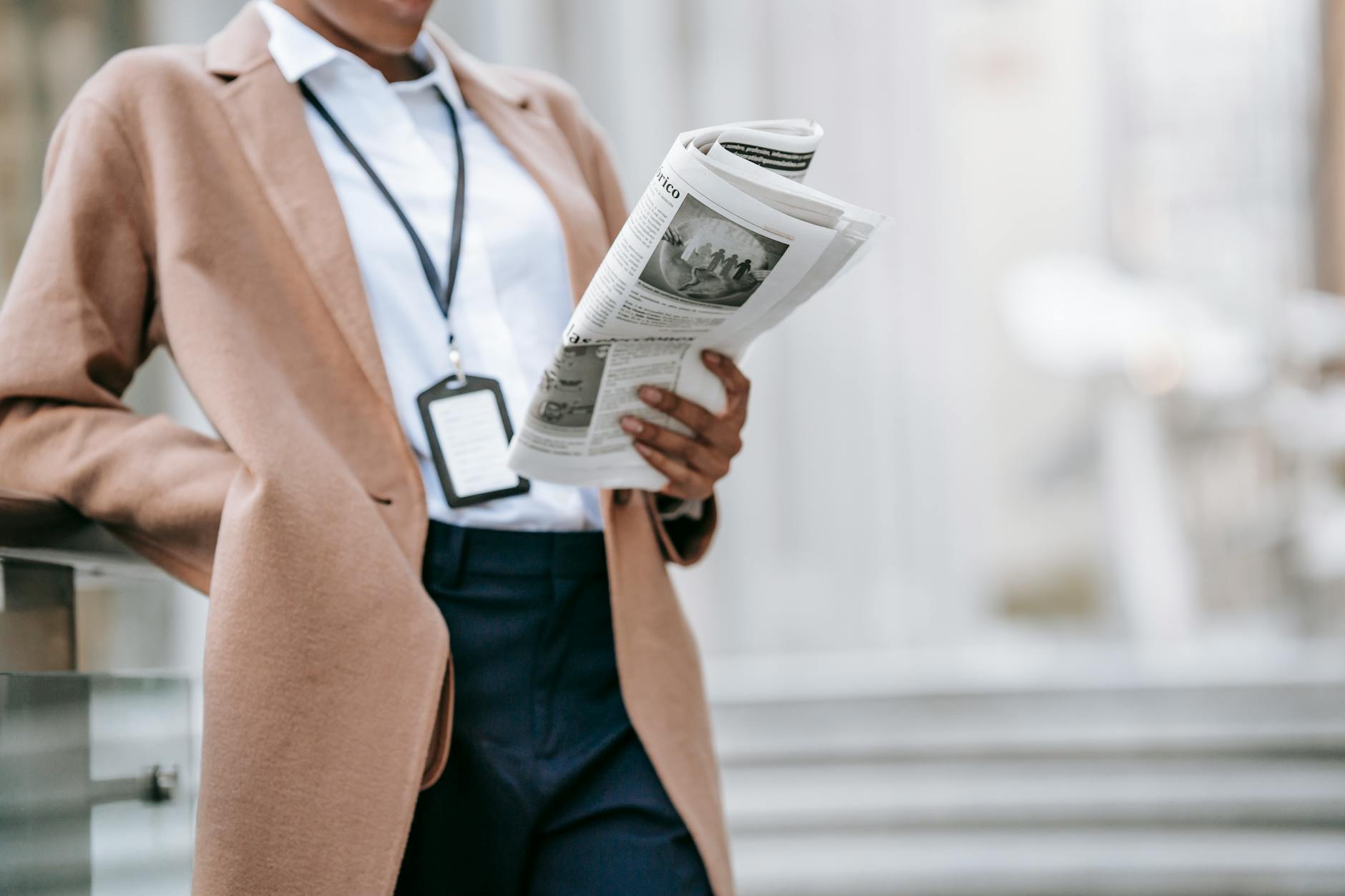 Business professional in camel coat and lanyard reading a folded newspaper outside an office building on urban steps