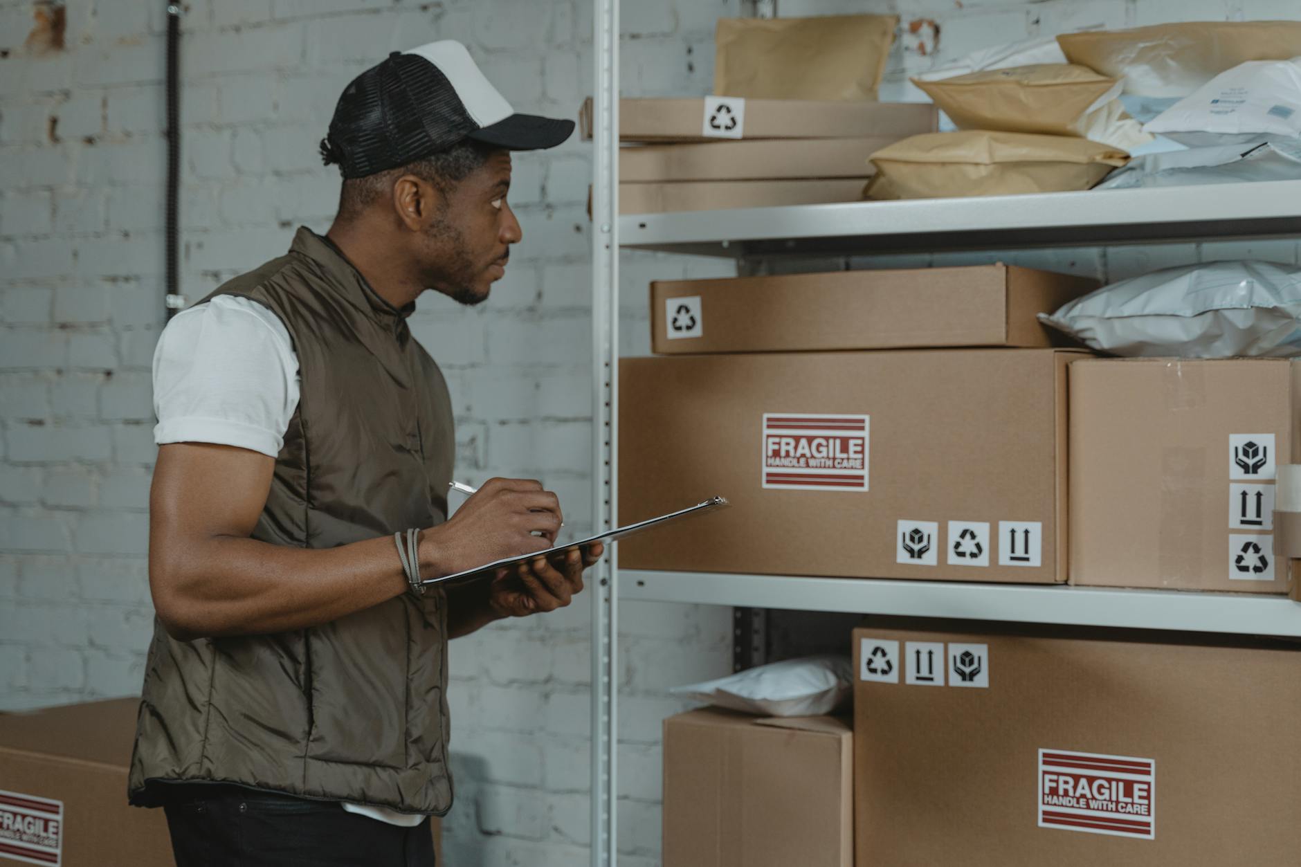 Warehouse worker checking inventory on a clipboard beside shelves of cardboard shipping boxes labeled "FRAGILE" and recycling symbols, organized storage room for e-commerce fulfillment and fragile item packing.