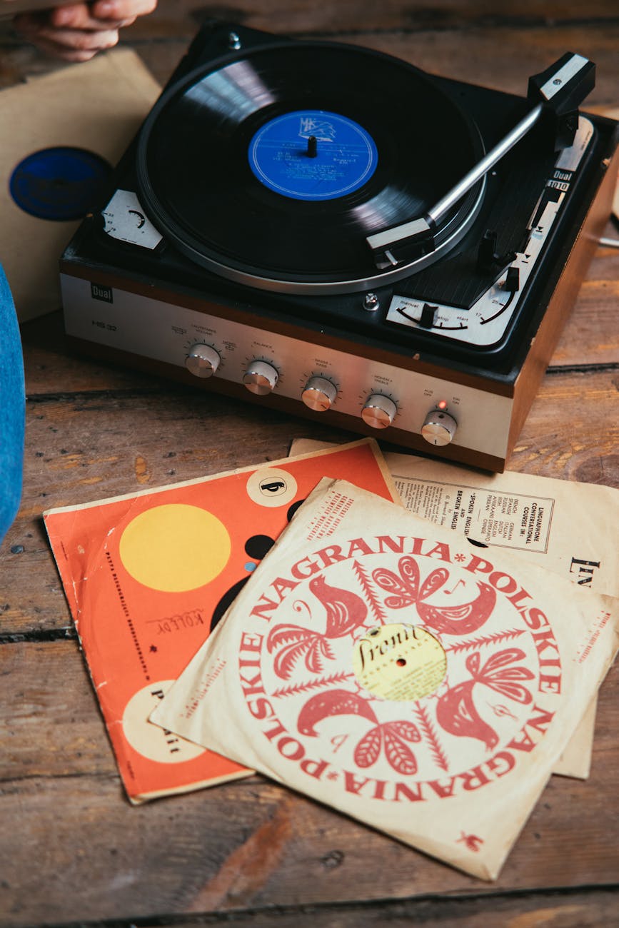 Vintage turntable playing a black vinyl record on a wooden table surrounded by retro album sleeves and colorful Polish record cover, close-up of record player controls and record sleeve artwork for music, retro audio, and vinyl enthusiasts.