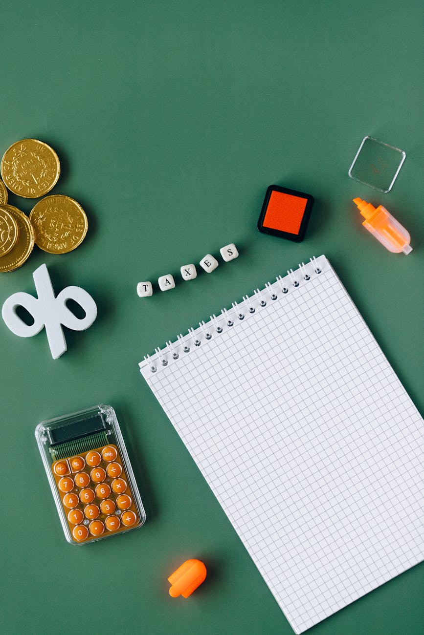 Flat lay of tax and finance items on green background: spiral graph notepad, orange calculator and highlighter, gold coins, white percent symbol and letter tiles spelling TAXES — concept image for taxes, accounting, budgeting