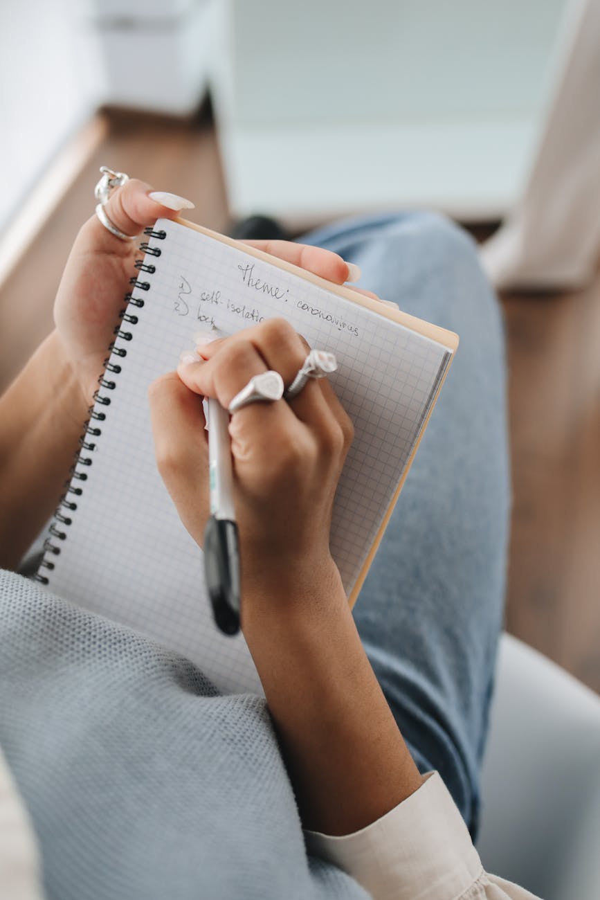 Close-up of hands with rings writing notes in a spiral grid notebook with a black pen, casual jeans and sweater in soft indoor light