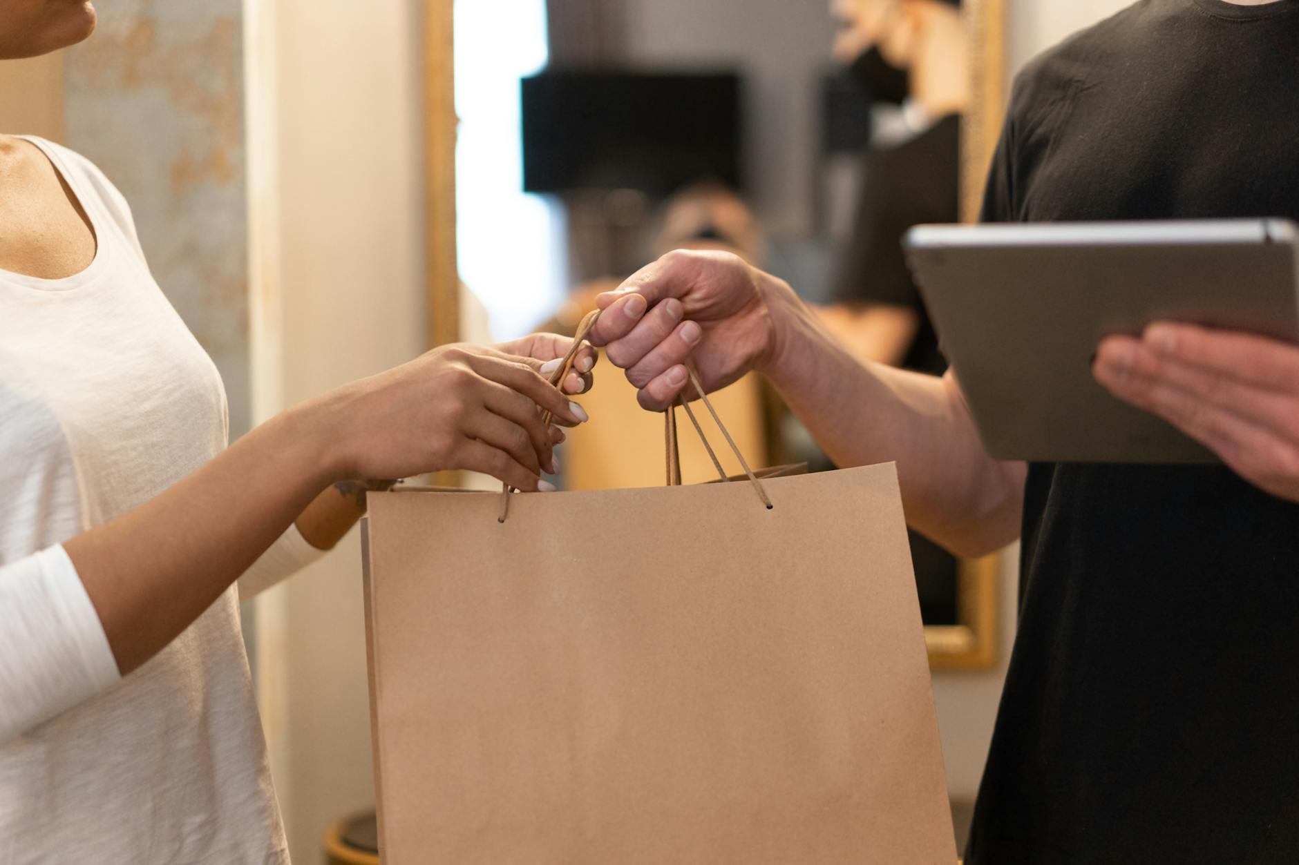 Close-up of a customer receiving a brown paper shopping bag from a delivery person holding a tablet, hands exchanging purchase in a retail setting