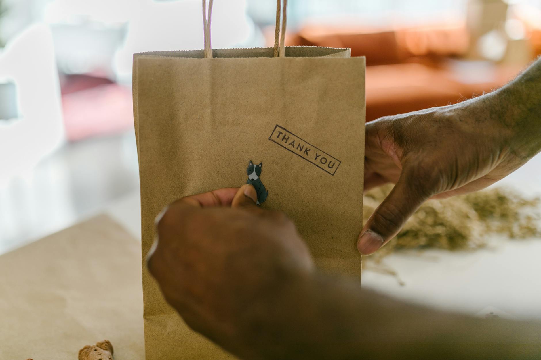 Hands attaching a small animal sticker to a brown paper gift bag stamped "THANK YOU" on a tabletop, eco-friendly packaging scene