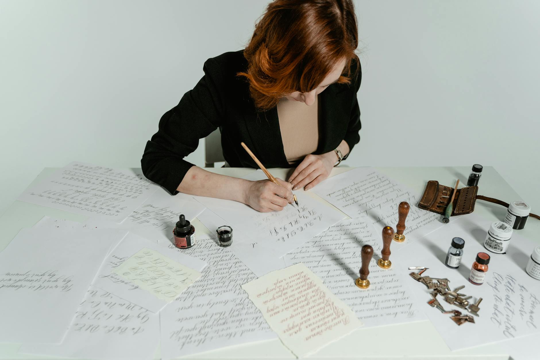 Woman practicing calligraphy at a white desk surrounded by handwritten papers, dip pens, ink bottles, wax seals and calligraphy tools, modern lettering workspace.