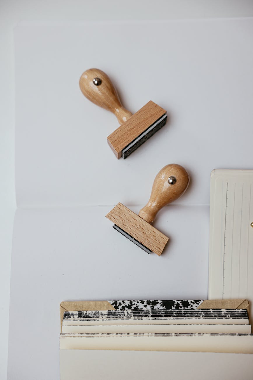 Two wooden rubber stamps on white paper beside a lined notebook and stacked journals — minimalist stationery scene for crafting, office supplies, journaling, and DIY stamping projects.