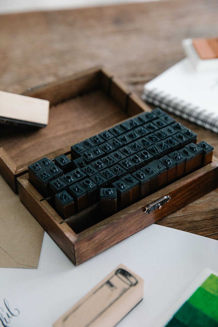 Wooden box filled with black rubber alphabet and symbol stamps on a rustic table, surrounded by a notebook, envelopes and craft stamps — DIY rubber stamp set for card making, scrapbooking, and personalized stationery.