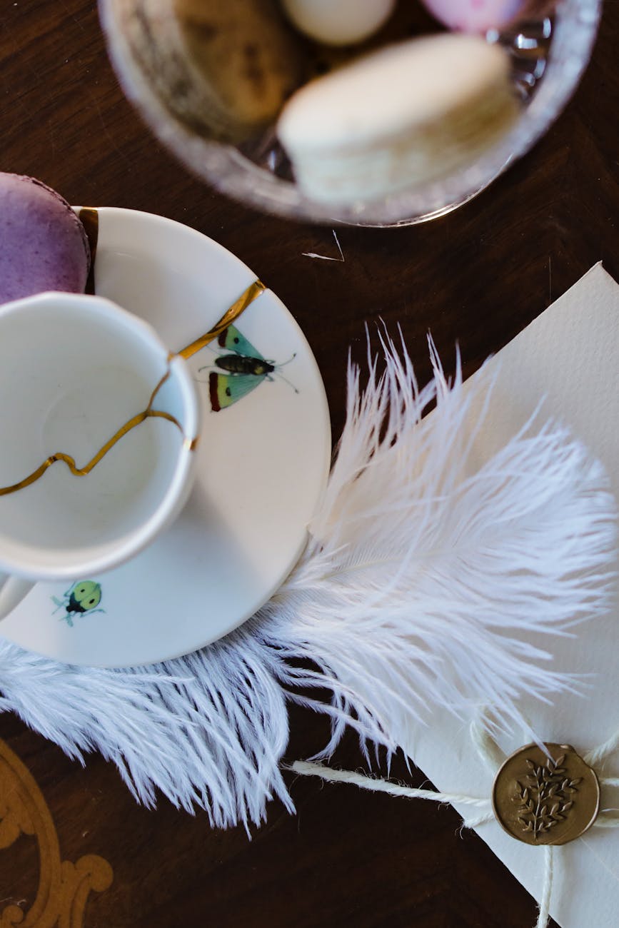 Flatlay of a kintsugi-repaired porcelain teacup and saucer with insect motif, white feather, wax-sealed envelope and pastel macarons on dark wood