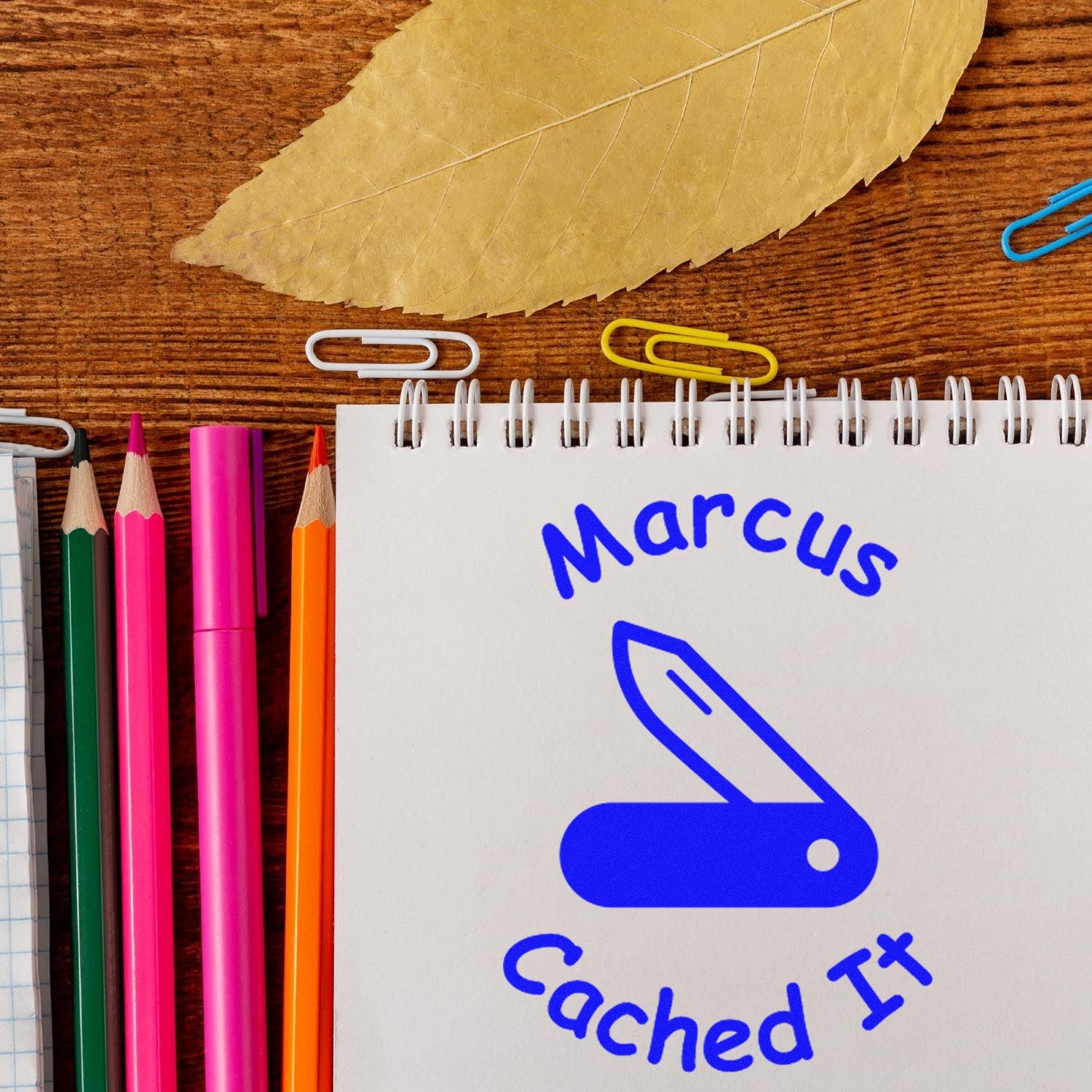 Blue Marcus Cached It self-inking stamp seal on a white spiral notebook, with colorful pencils and paper clips on wood.