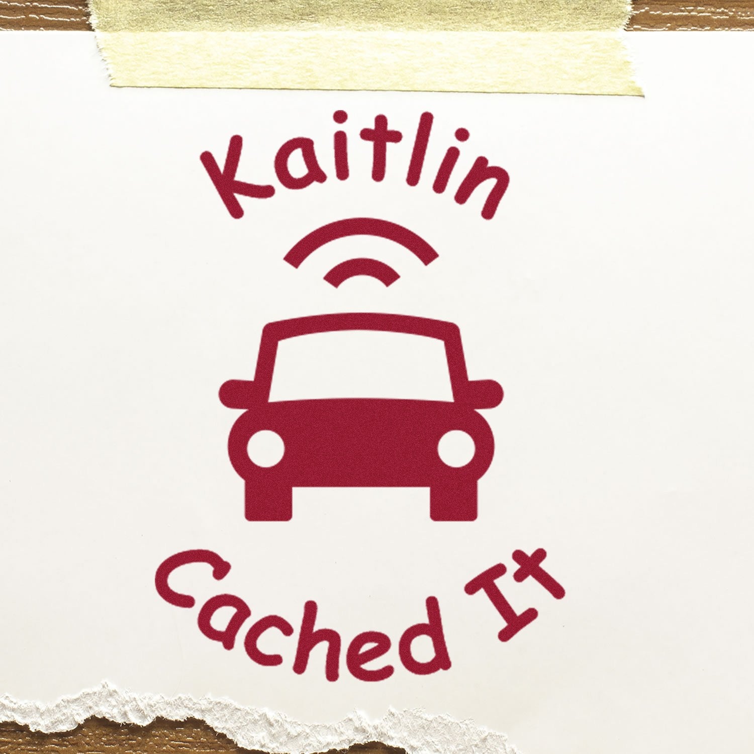 Burgundy Kaitlin Cached It stamp seal with car icon on white paper, taped to wood.