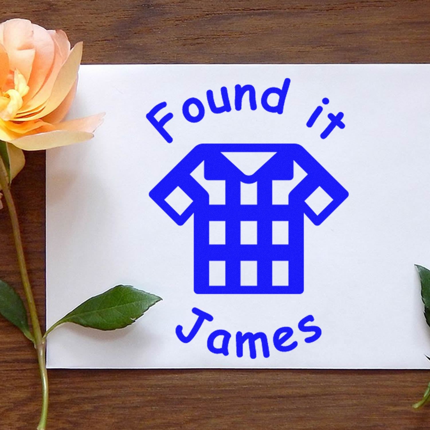Blue custom stamp and seal for clothing, reading Found it James, on white card with peach flower accents.
