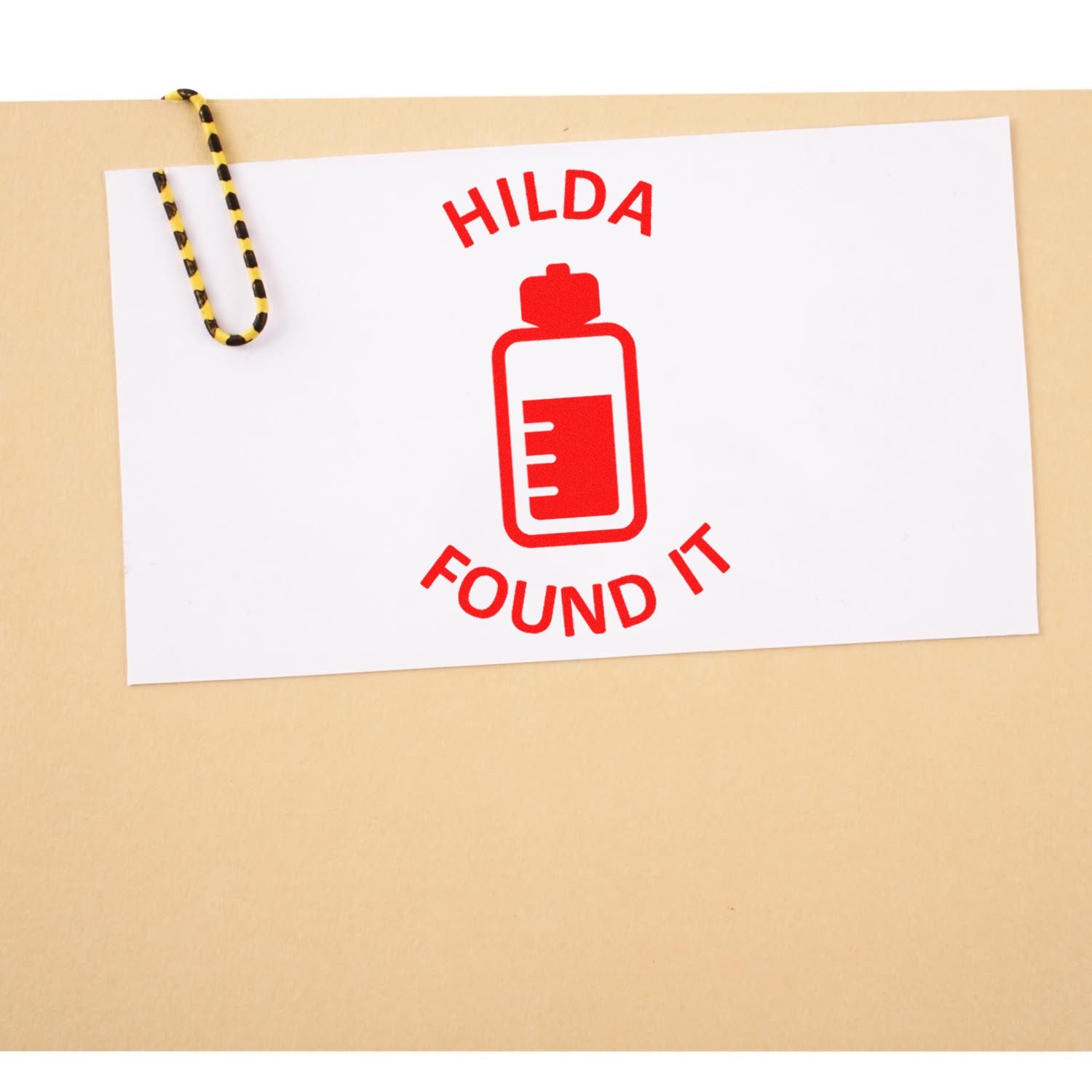 Red HILDA FOUND IT stamp and seal logo with bottle icon, clipped to beige paper by a yellow-black paperclip.
