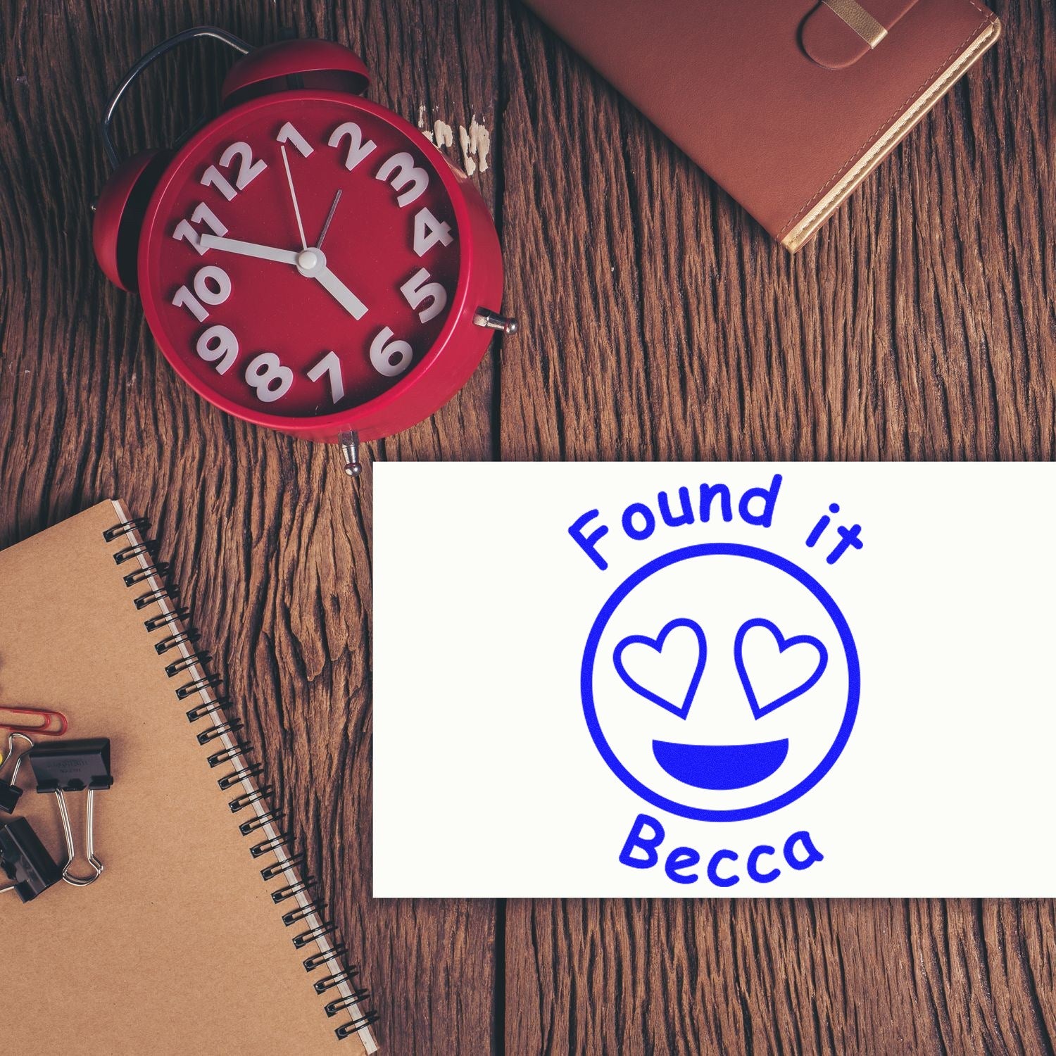 Blue heart-eyes custom stamp seal reading Found it Becca beside a red alarm clock on a wood desk.