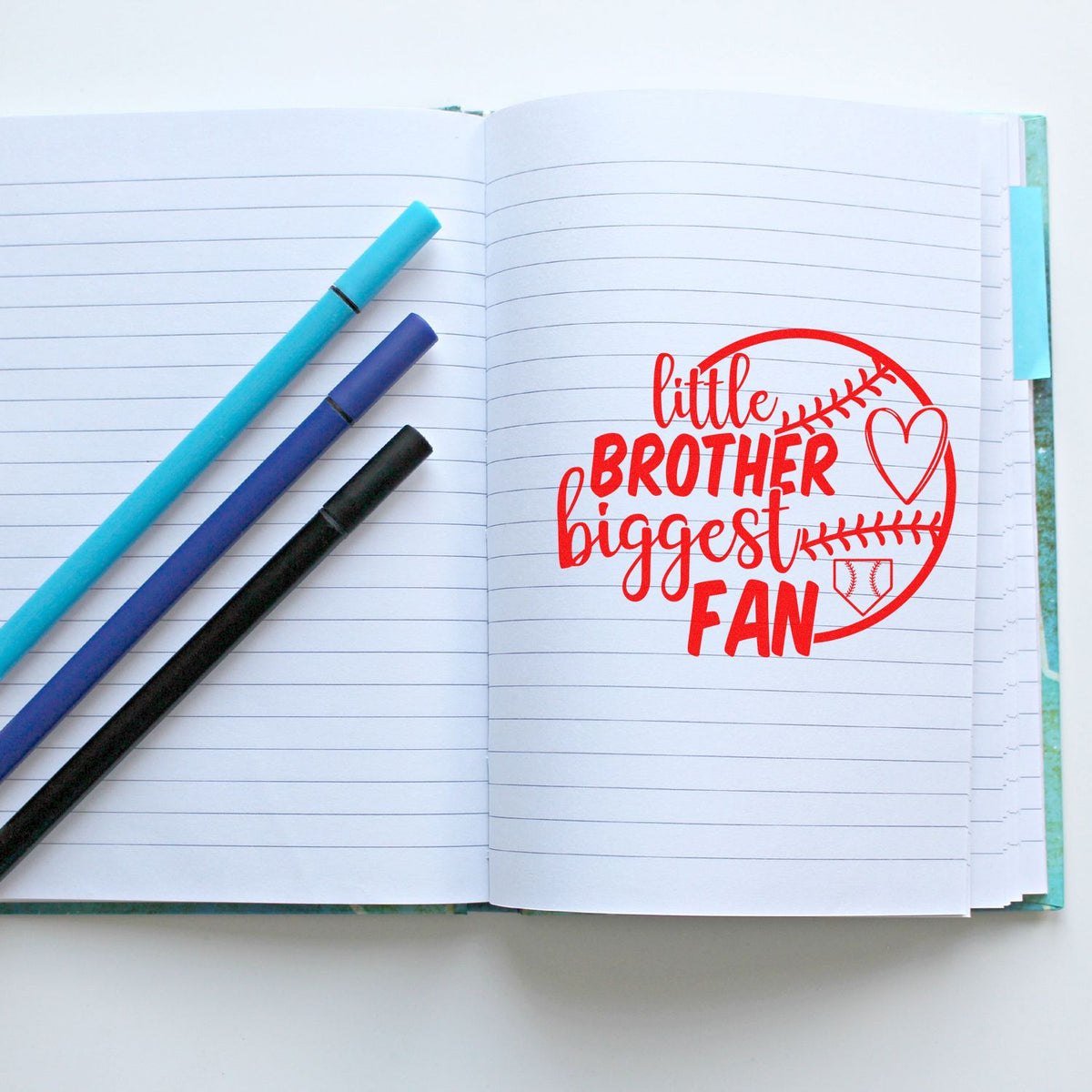 Red baseball-themed stamp seal reading little brother biggest fan on lined notebook with three pens in blue, purple and black.