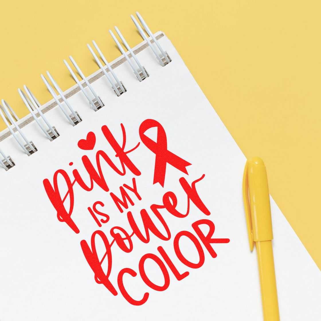 White spiral notebook with red breast cancer ribbon stamp reading Pink is my power color and a yellow pen on a yellow background.