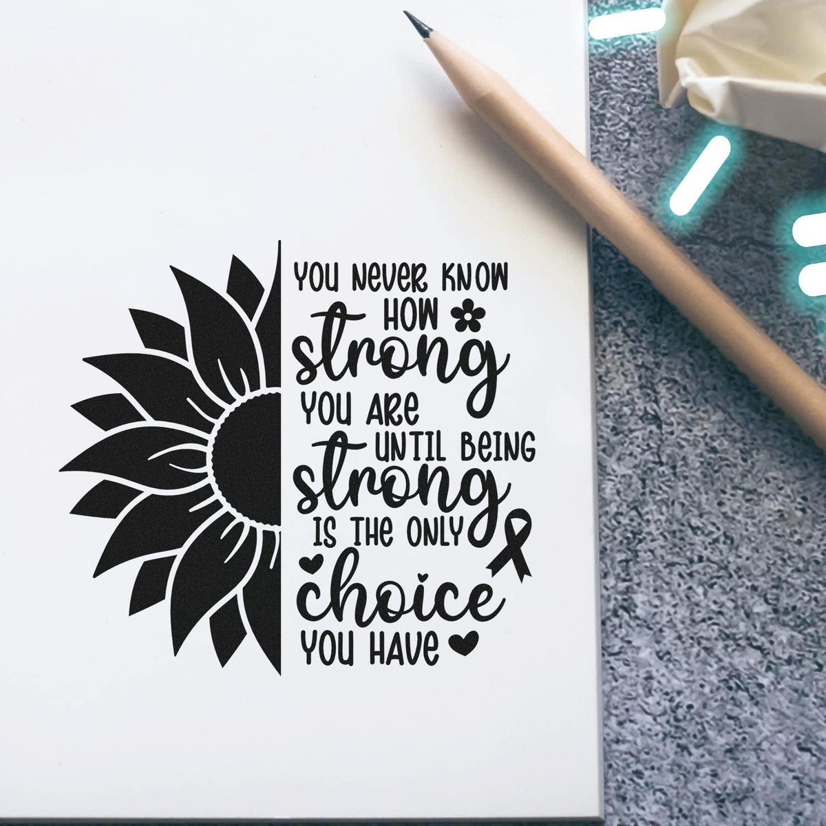 Black sunflower stamp and seal design with motivational quote on white paper beside a tan pencil.