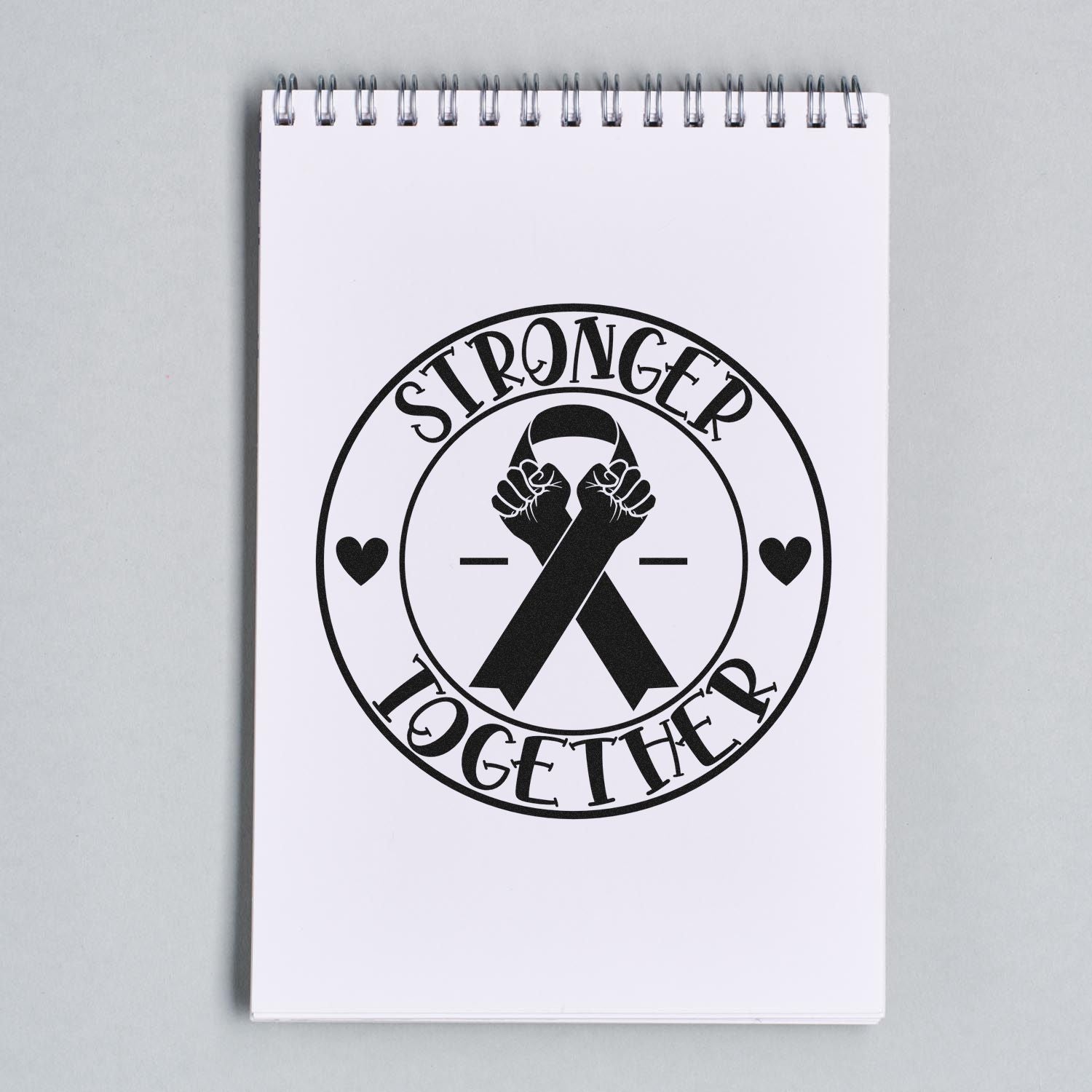 Black stamp seal on white spiral notebook reading Stronger Together, with raised fists, ribbons and hearts.