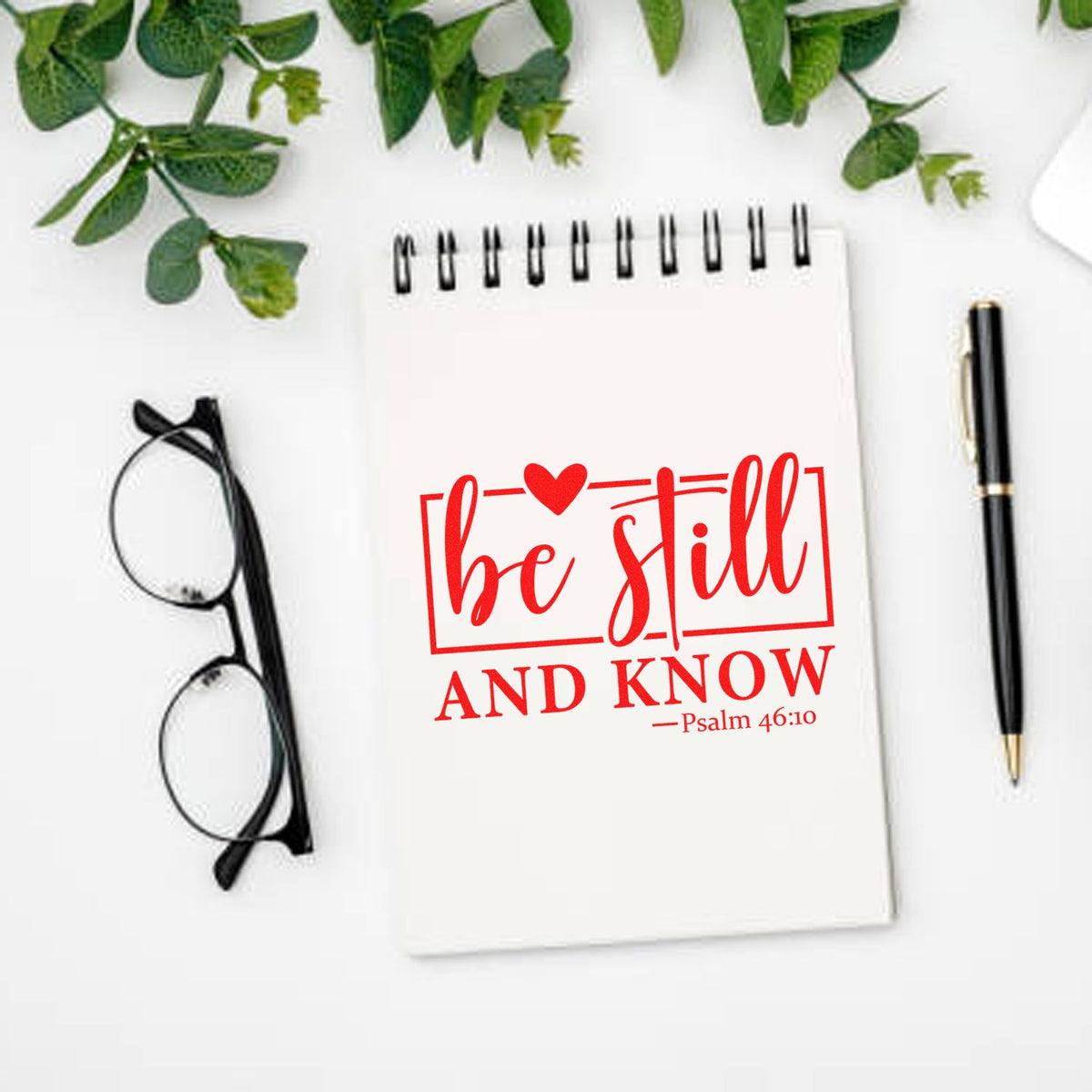 White notebook featuring a red custom stamp seal reading be still and know, with black glasses, pen, and green leaves