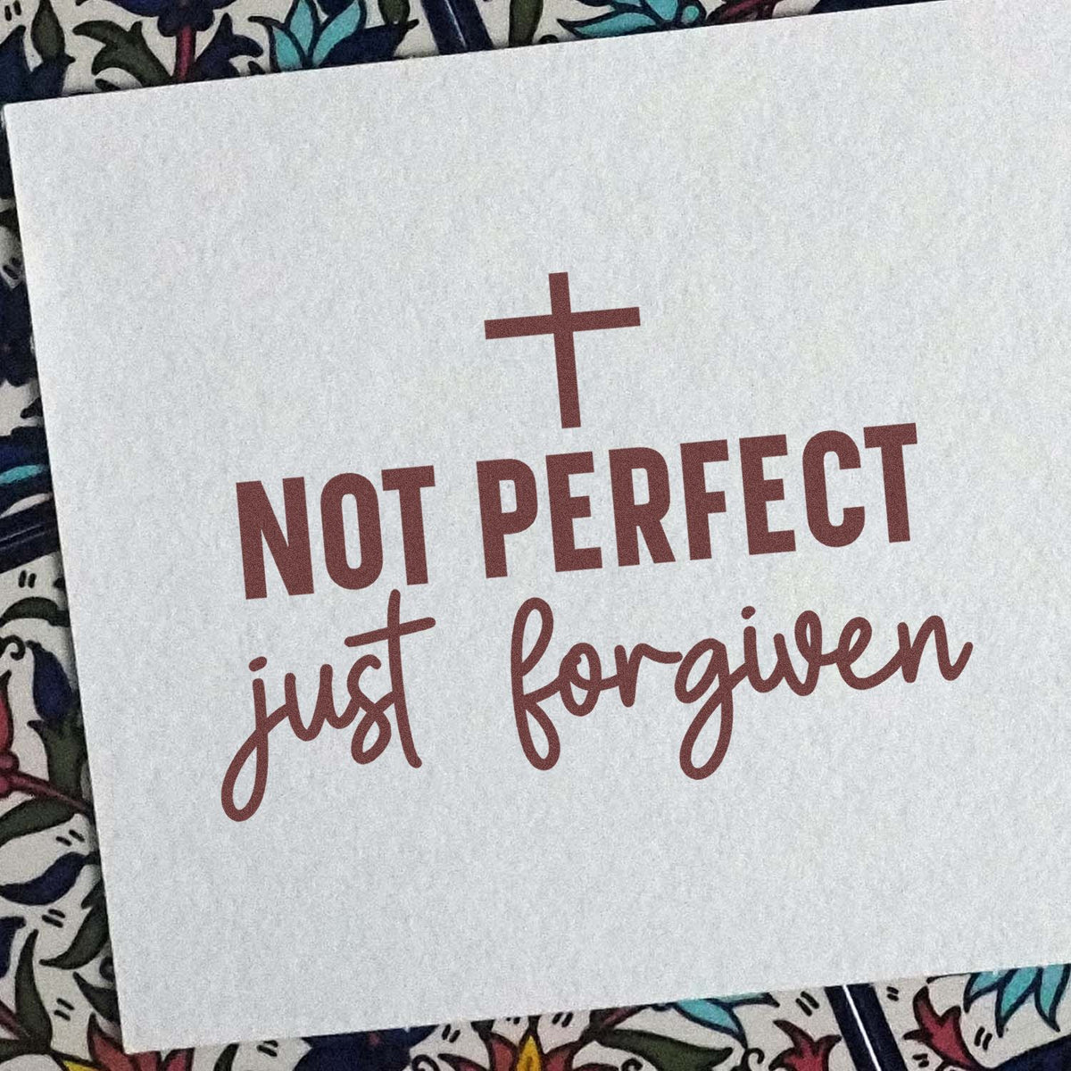 Maroon stamp and seal reading Not Perfect, Just Forgiven with a cross on a white background.