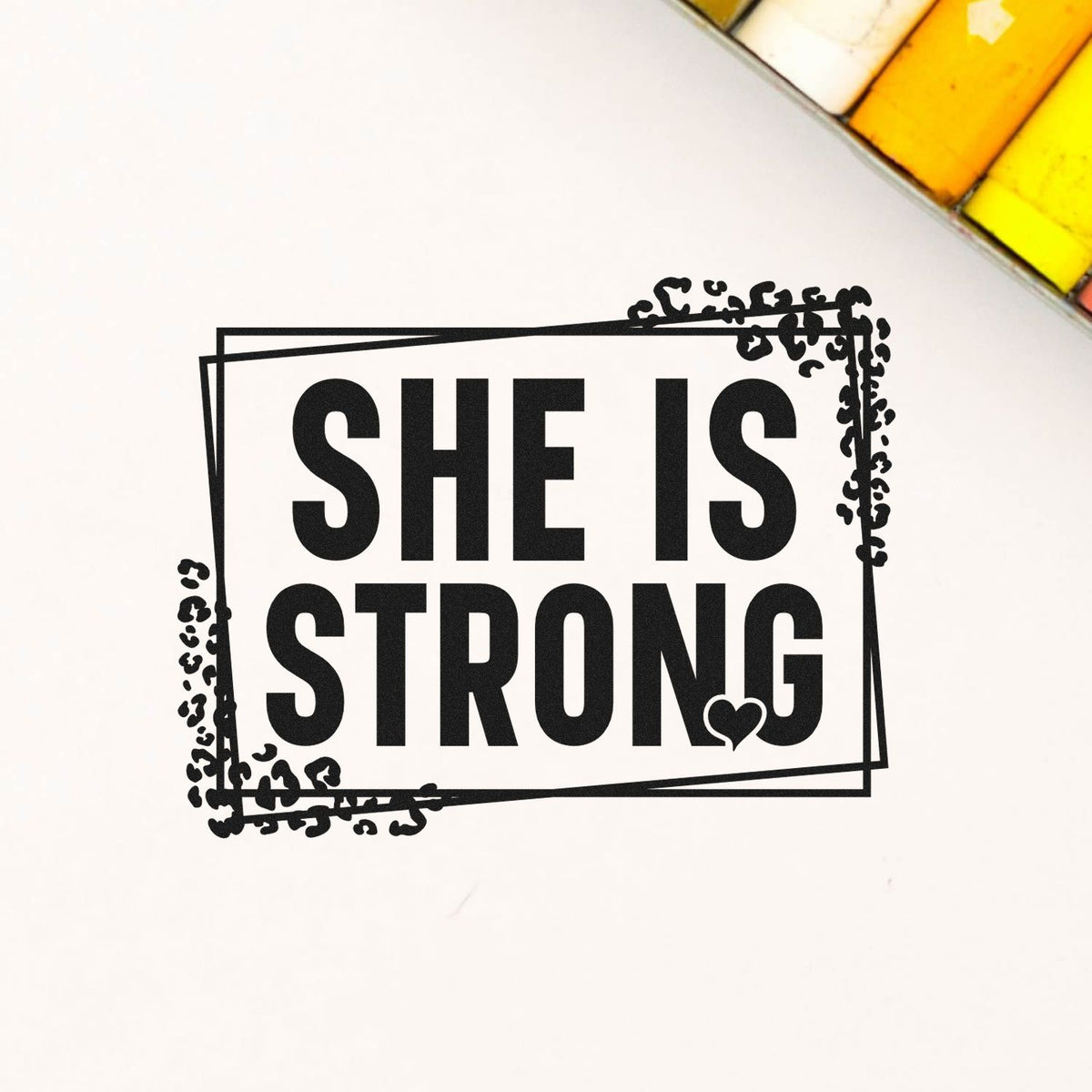 Black "She Is Strong" stamp seal with grunge border and heart on white background, accented by yellow corners.