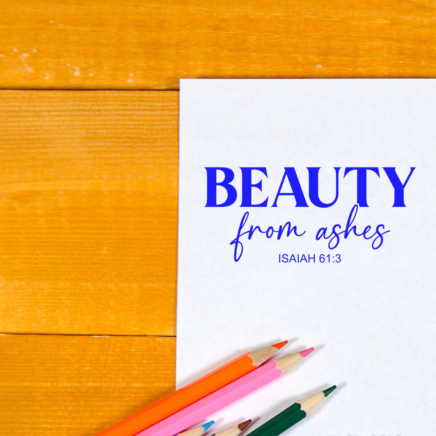 Blue stamp-style design on white paper reading Beauty from ashes, Isaiah 61:3, with colored pencils on honey wood.