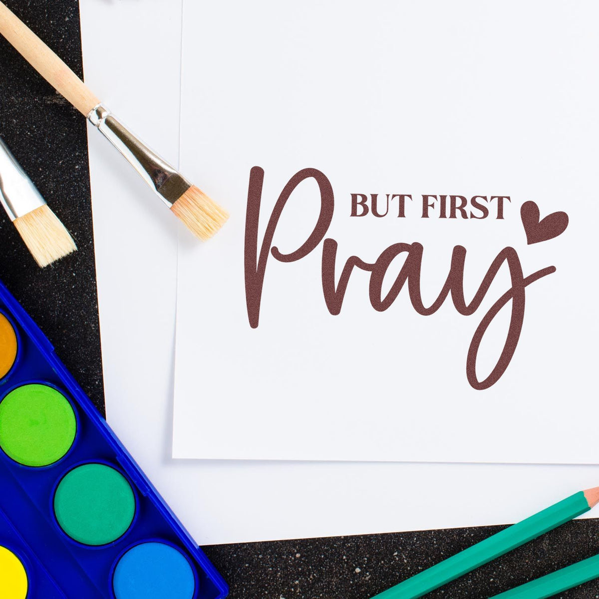 Brown But first Pray stamp seal on white paper, with heart, paintbrushes, watercolor palette, and teal pencils.