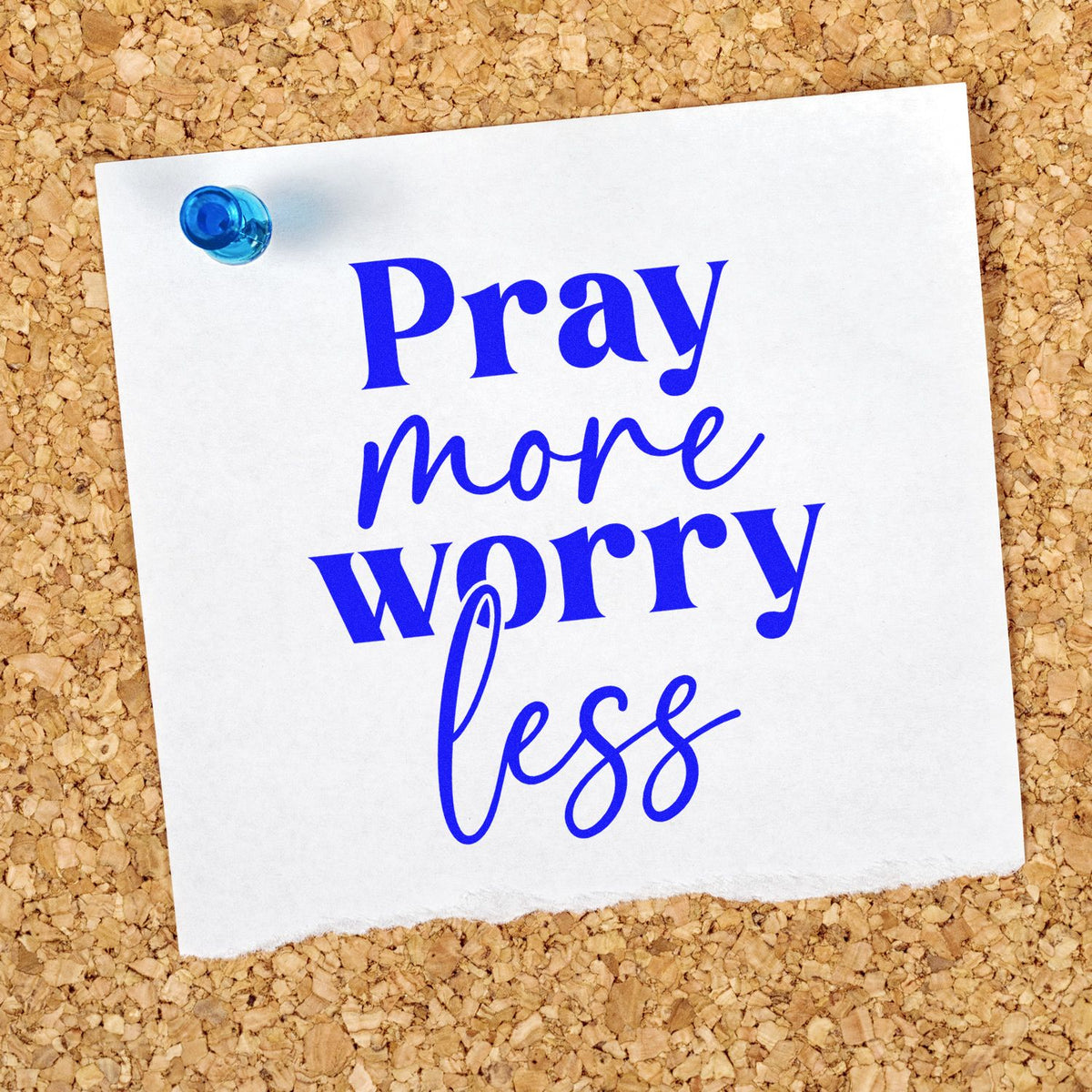 Blue rubber stamp reading Pray more worry less on white paper, pinned with a blue pushpin on a corkboard.