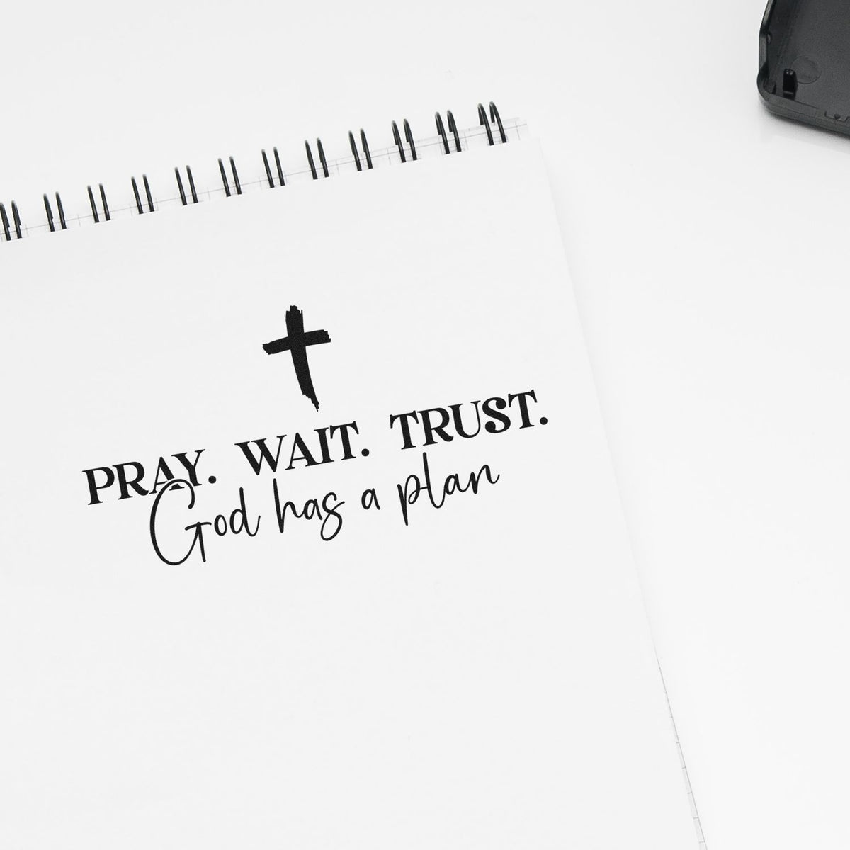 White spiral notebook featuring a black cross stamp and seal design with PRAY. WAIT. TRUST. God has a plan text.