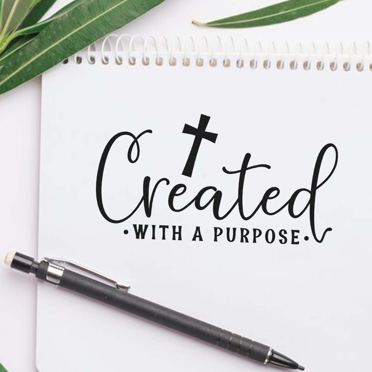 Black custom stamp seal reading Created with a purpose on a white notebook, with green leaves and a black pen.