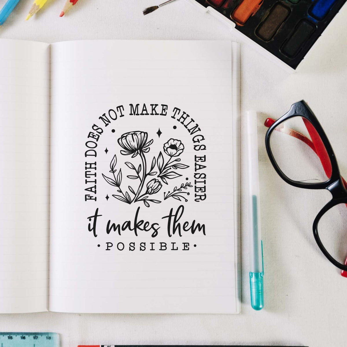 Black floral rubber stamp in open notebook, with black quote text, colorful pens, glasses, and watercolor palette on white desk.
