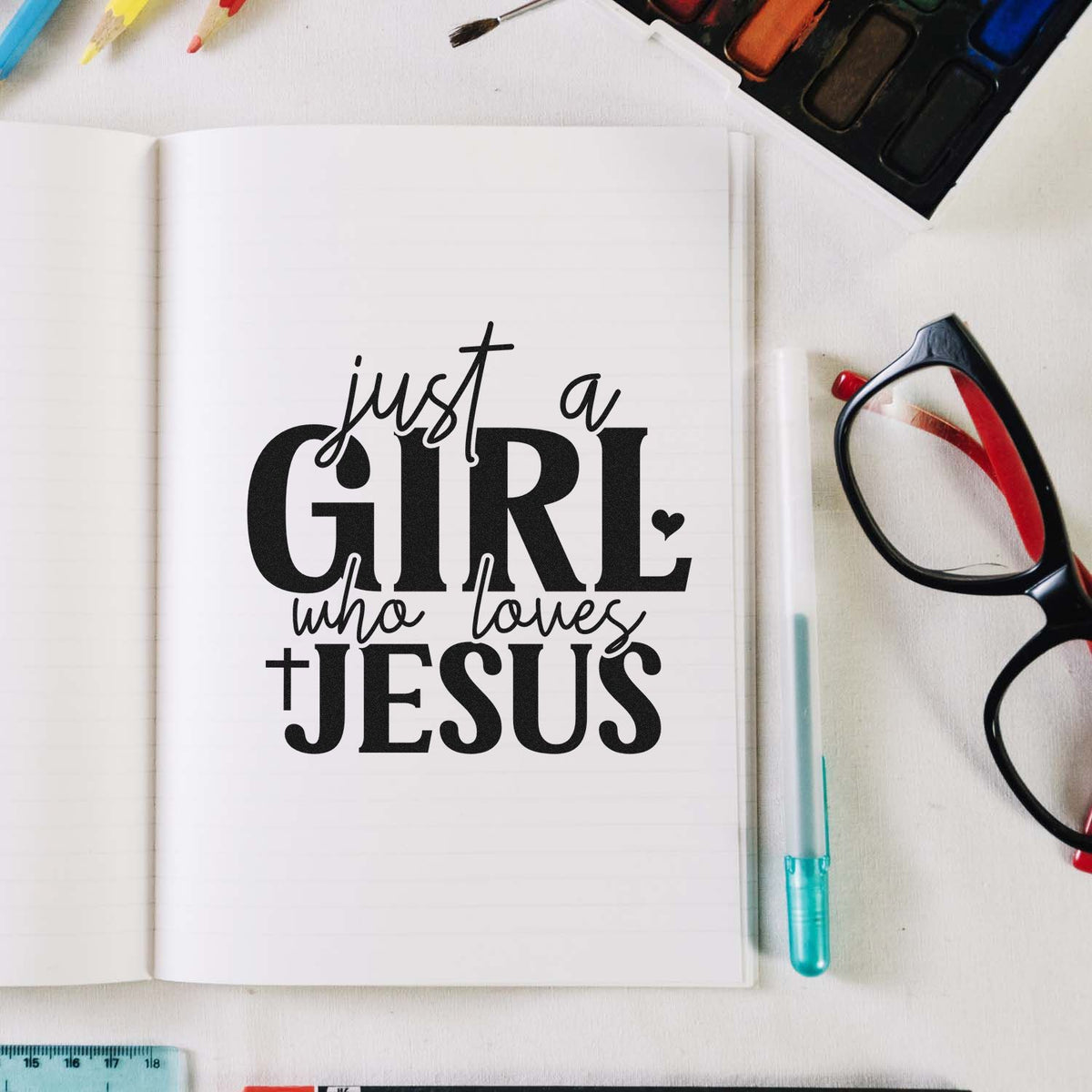Black stamp seal reading just a GIRL who loves JESUS on a white notebook, with paint palette, glasses, pen, and ruler nearby.