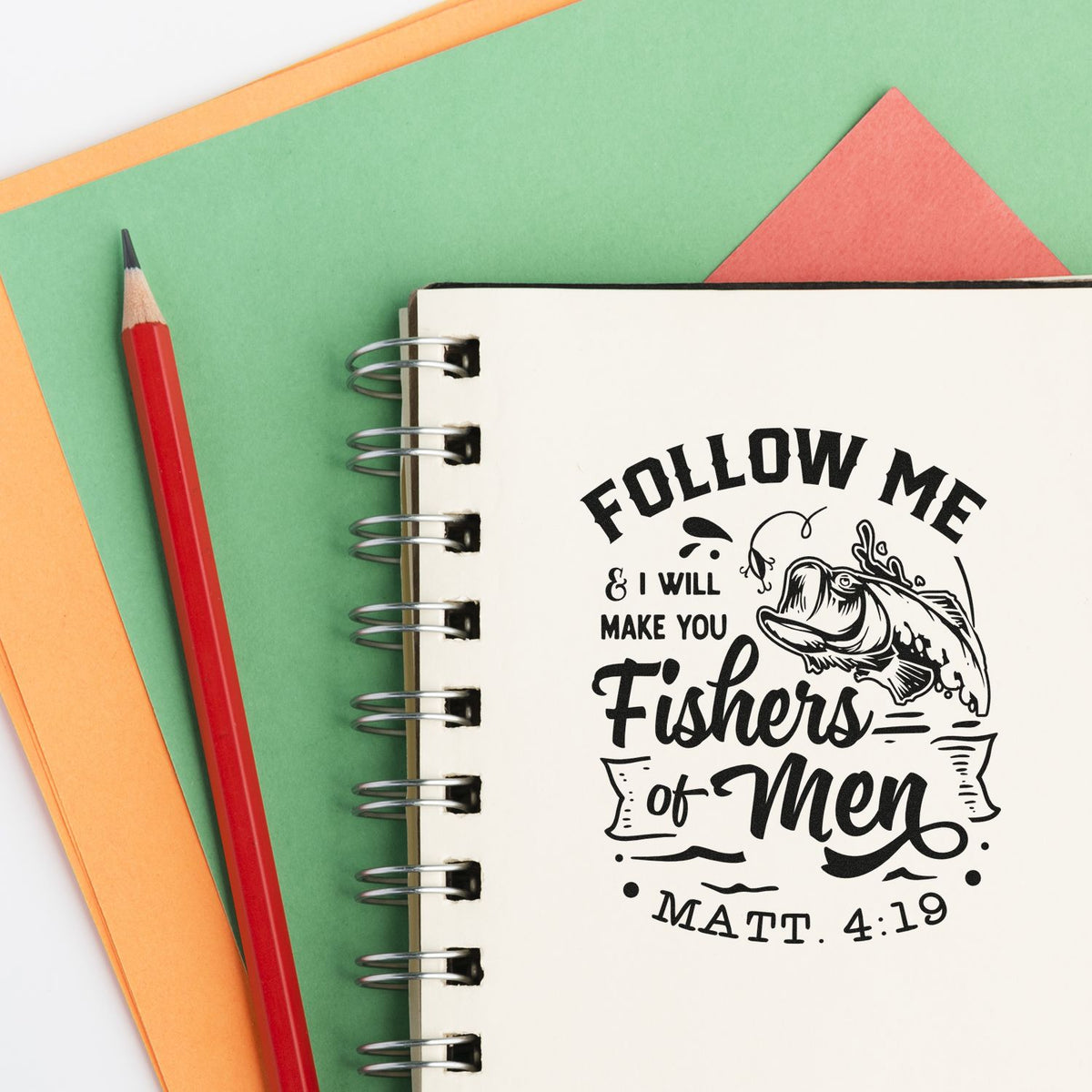 Black scripture stamp/seal on spiral notebook, with fish graphic, red pencil, and green-orange paper.