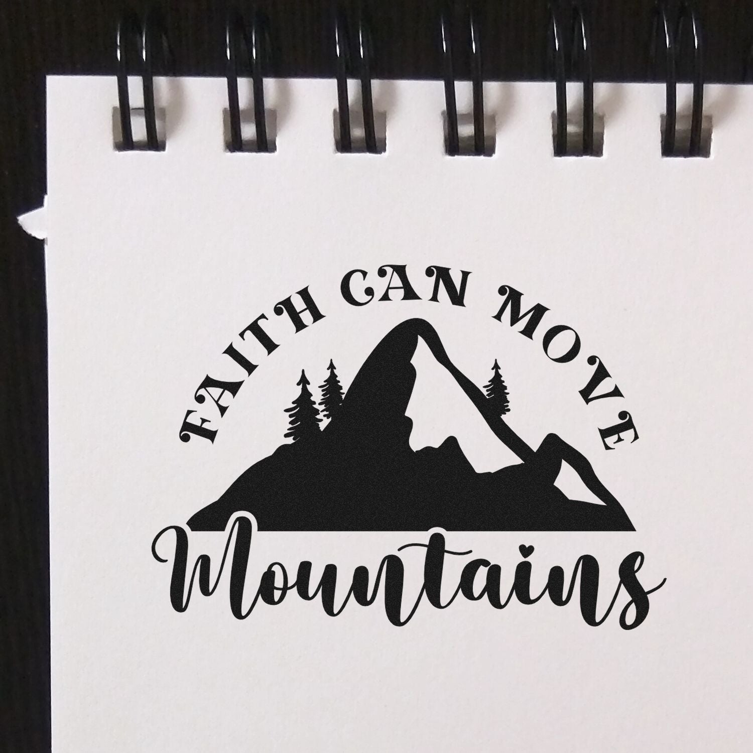Black Faith Can Move Mountains black stamp/seal design with mountain and pine trees on white paper in a spiral notebook.