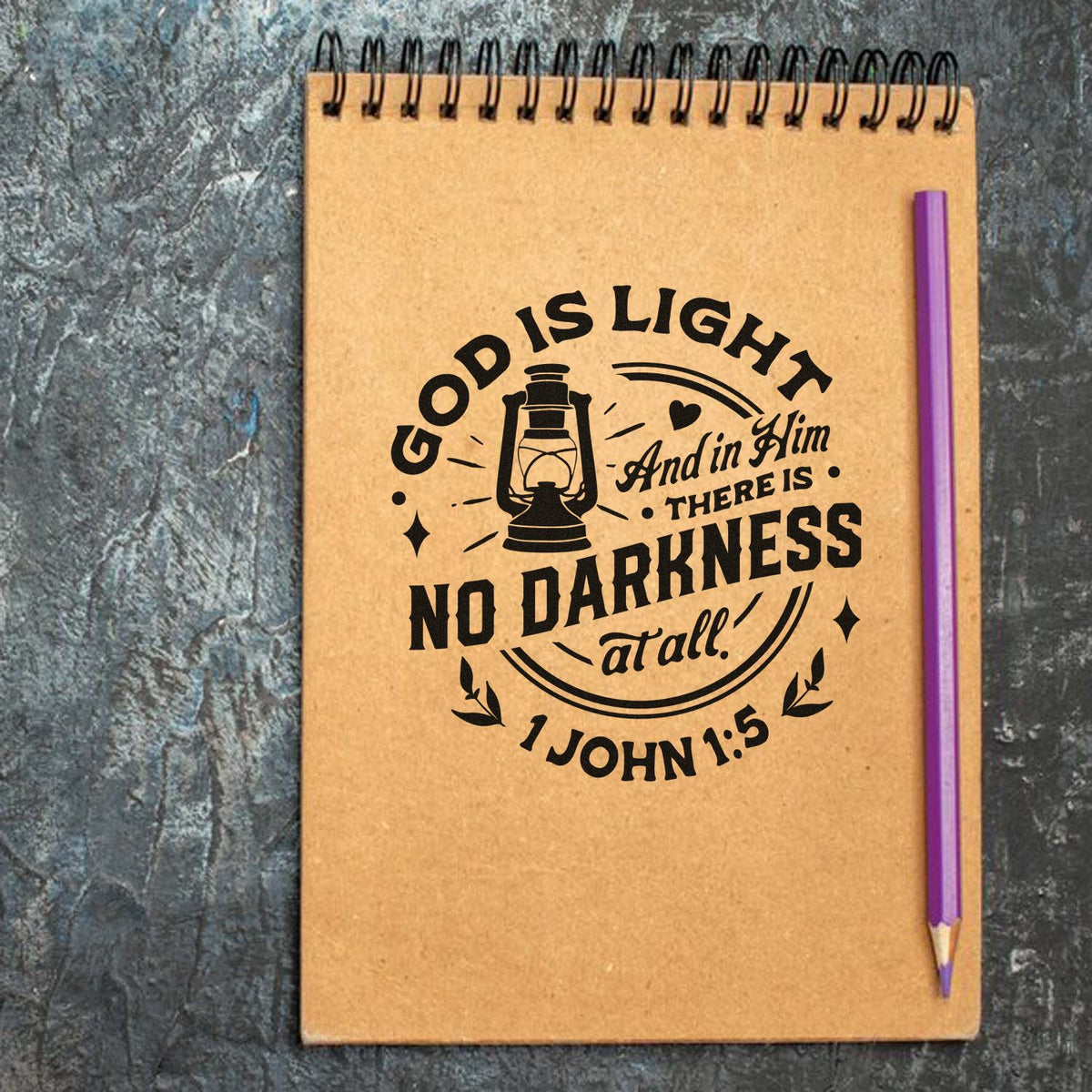 Brown spiral notebook featuring a black stamp seal with lantern and Bible verse, plus a purple pencil on a gray background.