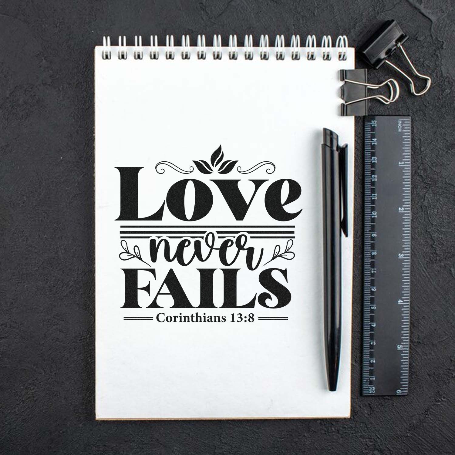 Black stamp seal design on white notebook: Love never fails with Corinthians 13:8, beside pen, ruler, and binder clip.